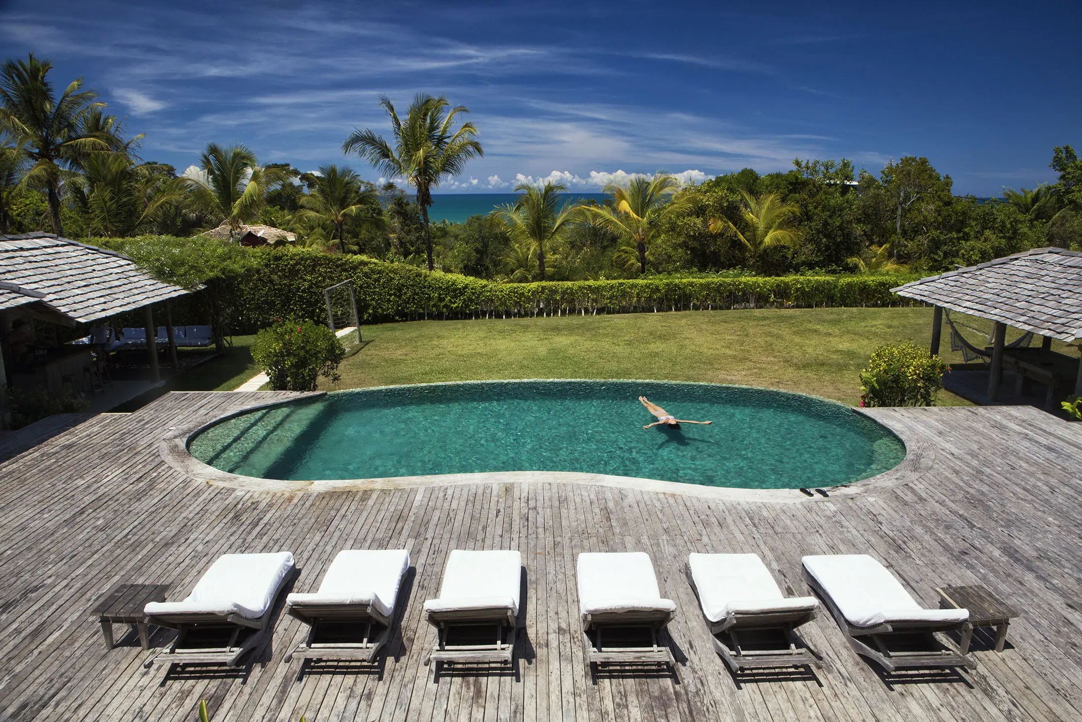 A serene outdoor scene featuring a kidney-shaped pool surrounded by a wooden deck with six white lounge chairs. One person relaxes in the pool. Lush greenery and palm trees frame the view, with a glimpse of the ocean and a clear blue sky in the background.