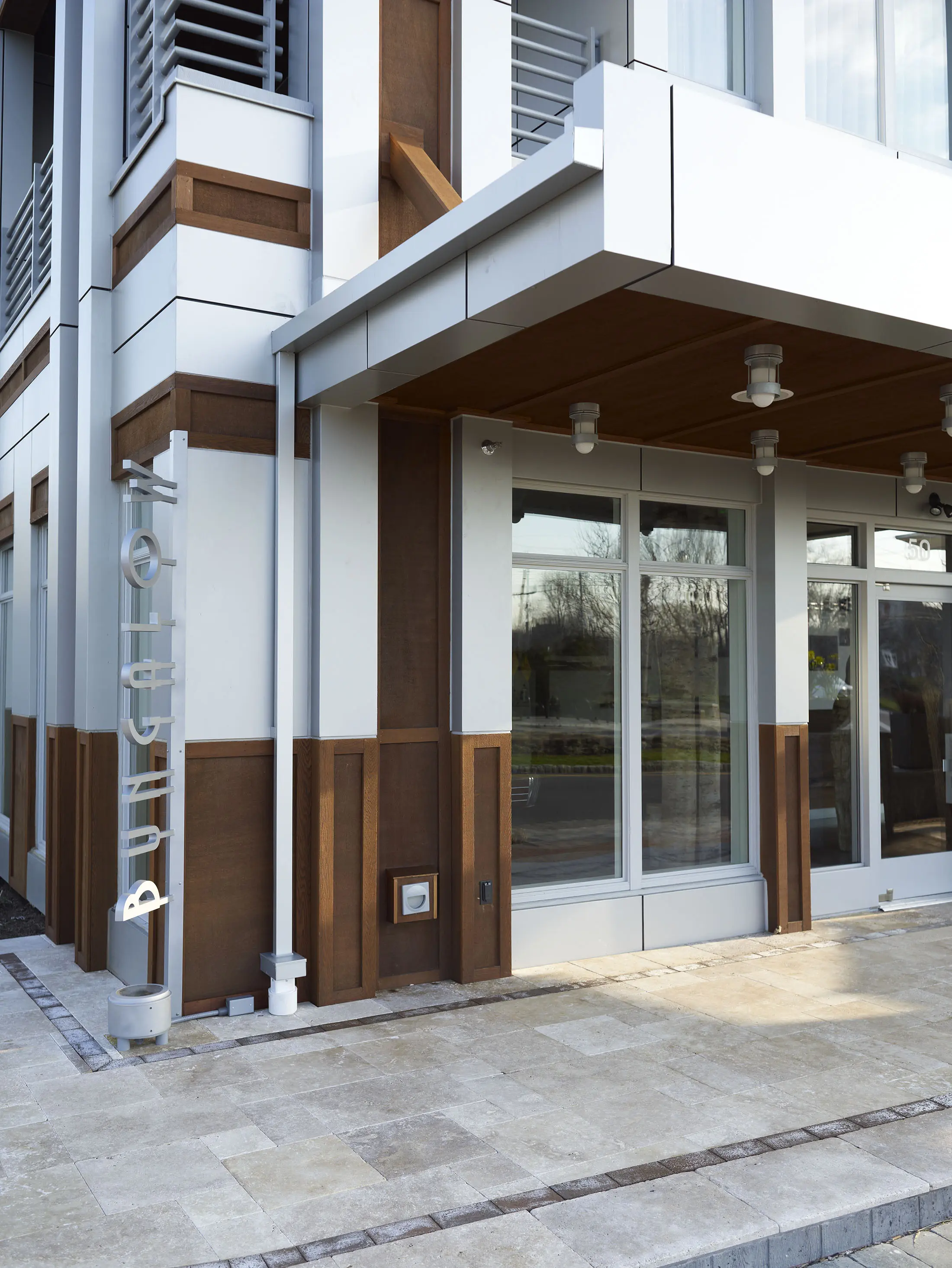 Modern building facade with a combination of white and brown panels. Large windows reflect a cloudy sky. The entrance features an awning with lights underneath. A vertical sign reads bungalow. The floor is paved with rectangular tiles, leading to a glass door.