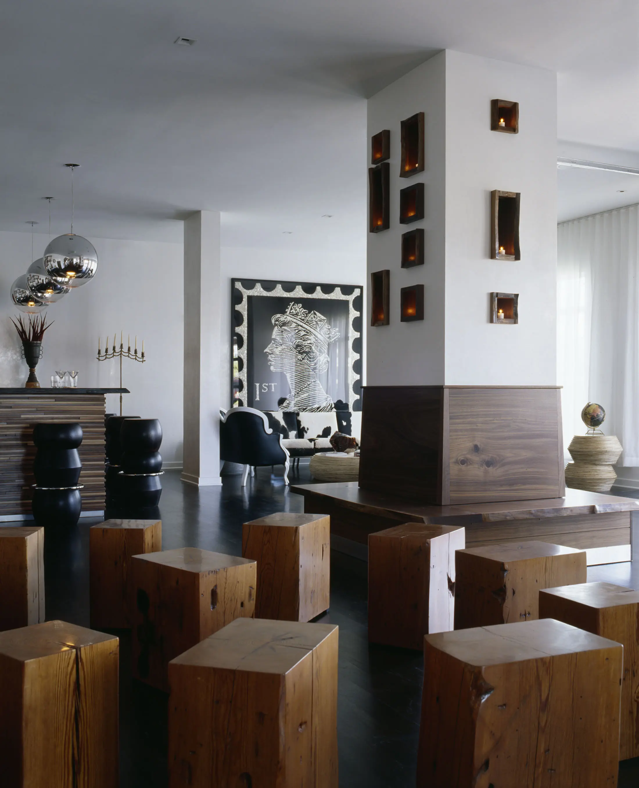 Contemporary interior with wooden block stools on dark flooring, a central white pillar with wooden niches and candles, a modern art piece featuring a womans profile on the wall, metallic spherical lamps, and white drapes. Mix of wooden and upholstered furniture completes the decor.