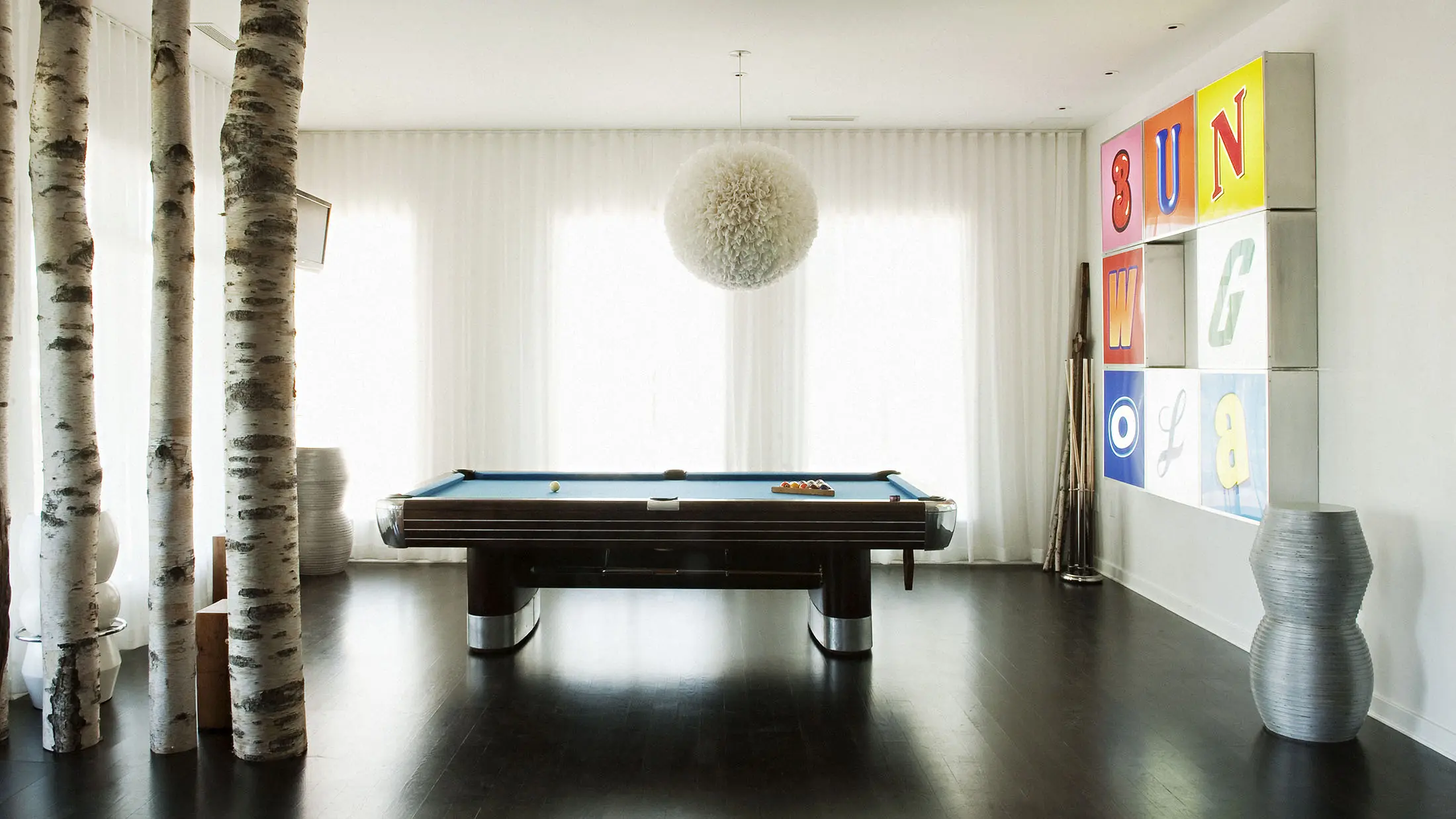 A modern room features a billiards table with blue felt in the center. Birch tree trunks stand to the left, while a fluffy pendant light hangs above. The right wall displays colorful, abstract art, and metallic vases are near the corner. Floor-to-ceiling white curtains cover the windows.