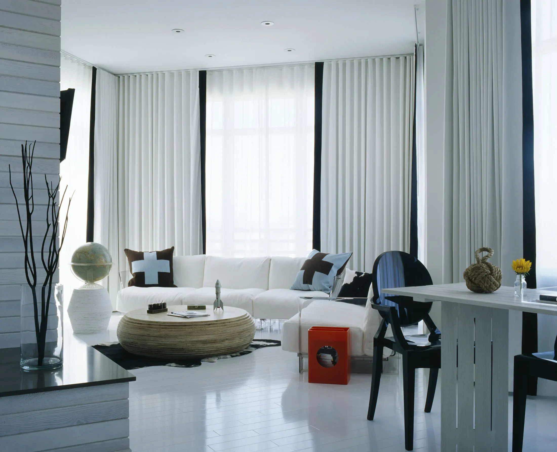A modern living room with white walls and floor, featuring a large white sectional sofa with black and white cushions. A round rattan coffee table holds books and decor. Black chairs, a globe, and a red side table accent the space. Floor-to-ceiling windows are covered with white curtains.