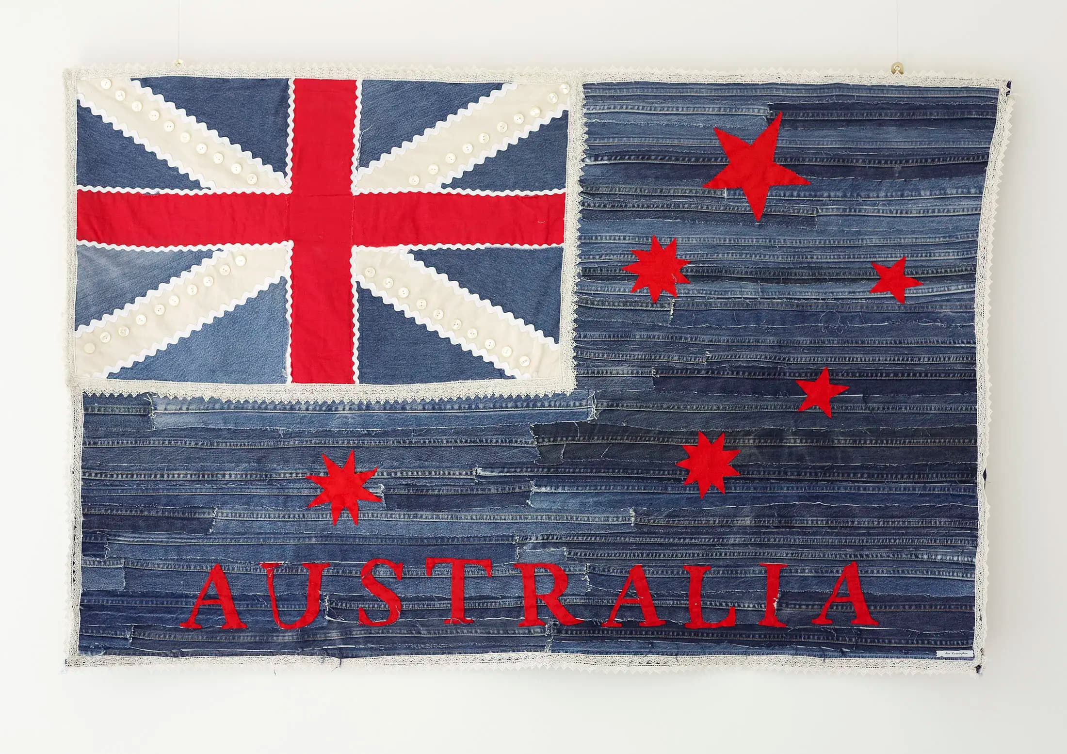 Patchwork textile art depicting an Australian flag. The Union Jack and Southern Cross are crafted from various denim shades and lace-like trim, with the word AUSTRALIA in red letters at the bottom. The background is textured with horizontal denim strips.
