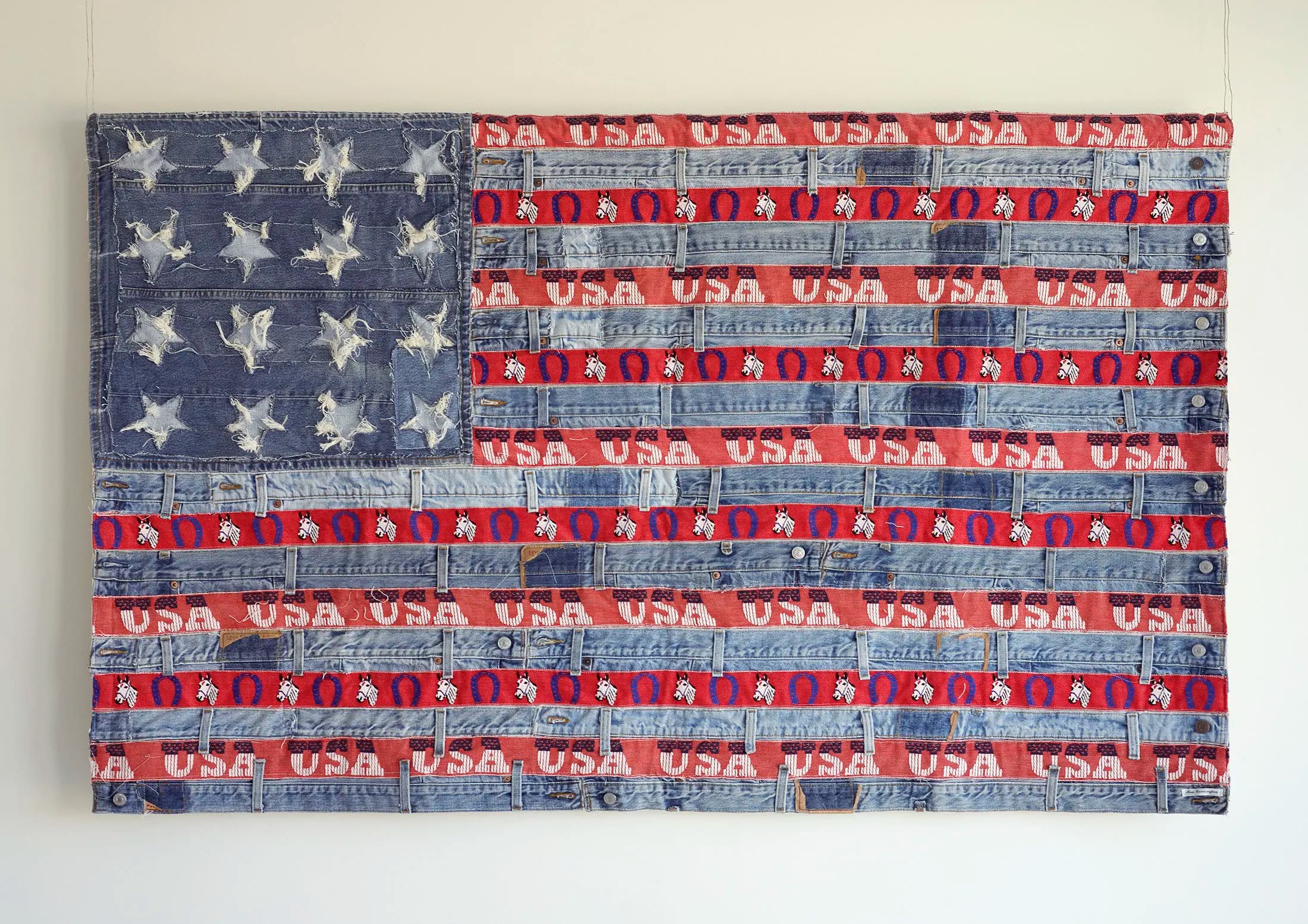 Artistic representation of the U.S. flag made from denim and red fabric strips. The canton features roughly cut denim stars on a blue denim background, while the stripes are interwoven with red fabric labeled USA and denim, presenting a textured, rustic appearance.