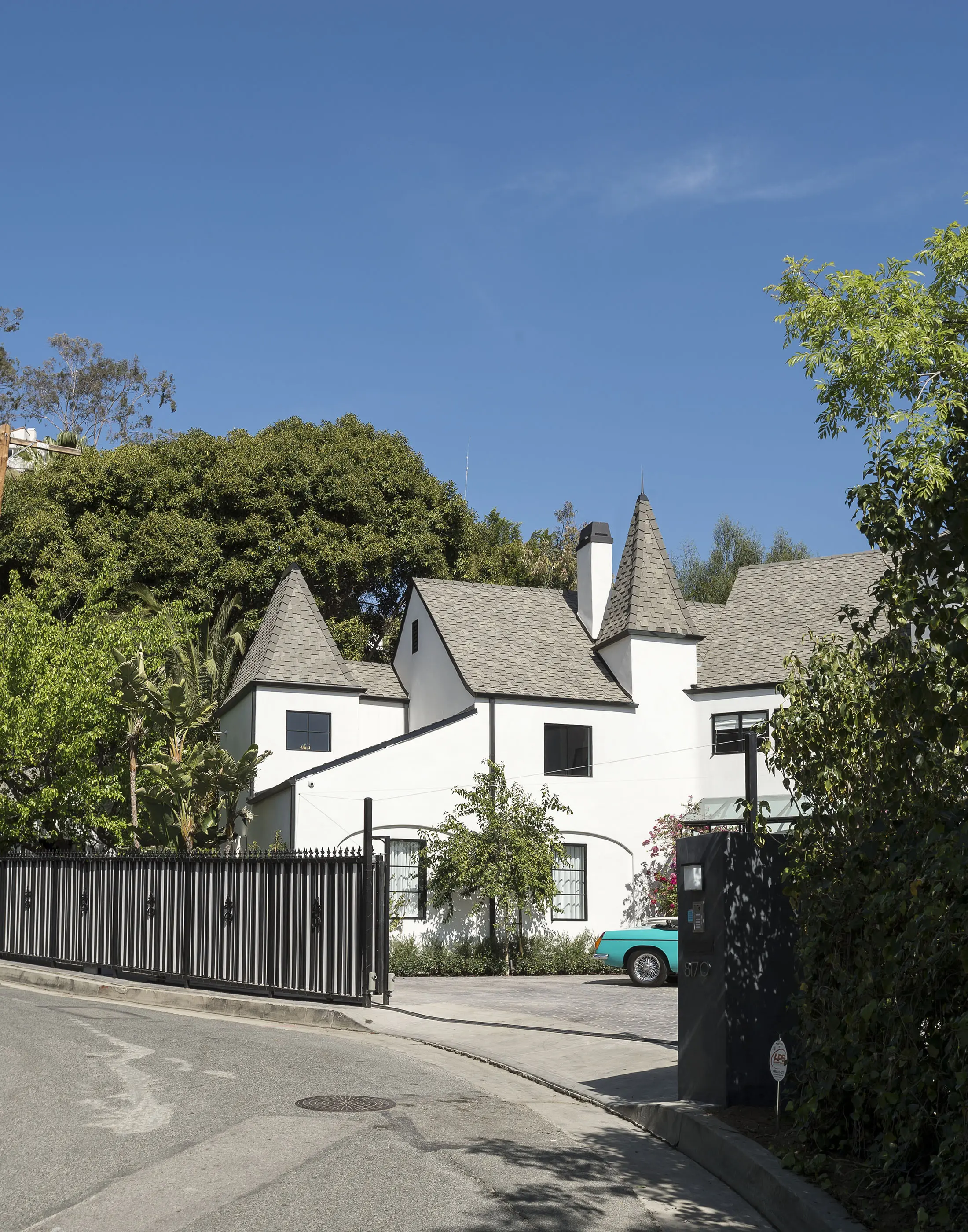 A white castle-like house with turrets and a steep gray roof, surrounded by greenery. It has a distinct black and white striped fence with ornate black ironwork along the top. The sky is clear and blue, and there is a tree partially visible on the left.