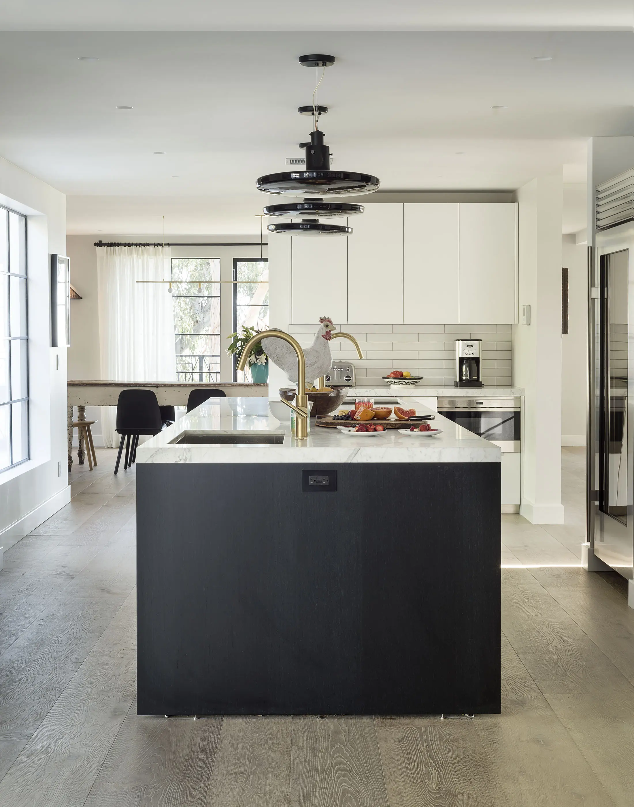 Modern kitchen with a large black island featuring a sink and gold faucet. White cabinets line the wall, and a stainless-steel fridge is visible. A round, tiered light fixture hangs above. Sunlight streams in through large windows, illuminating wooden floors and a dining area in the background.