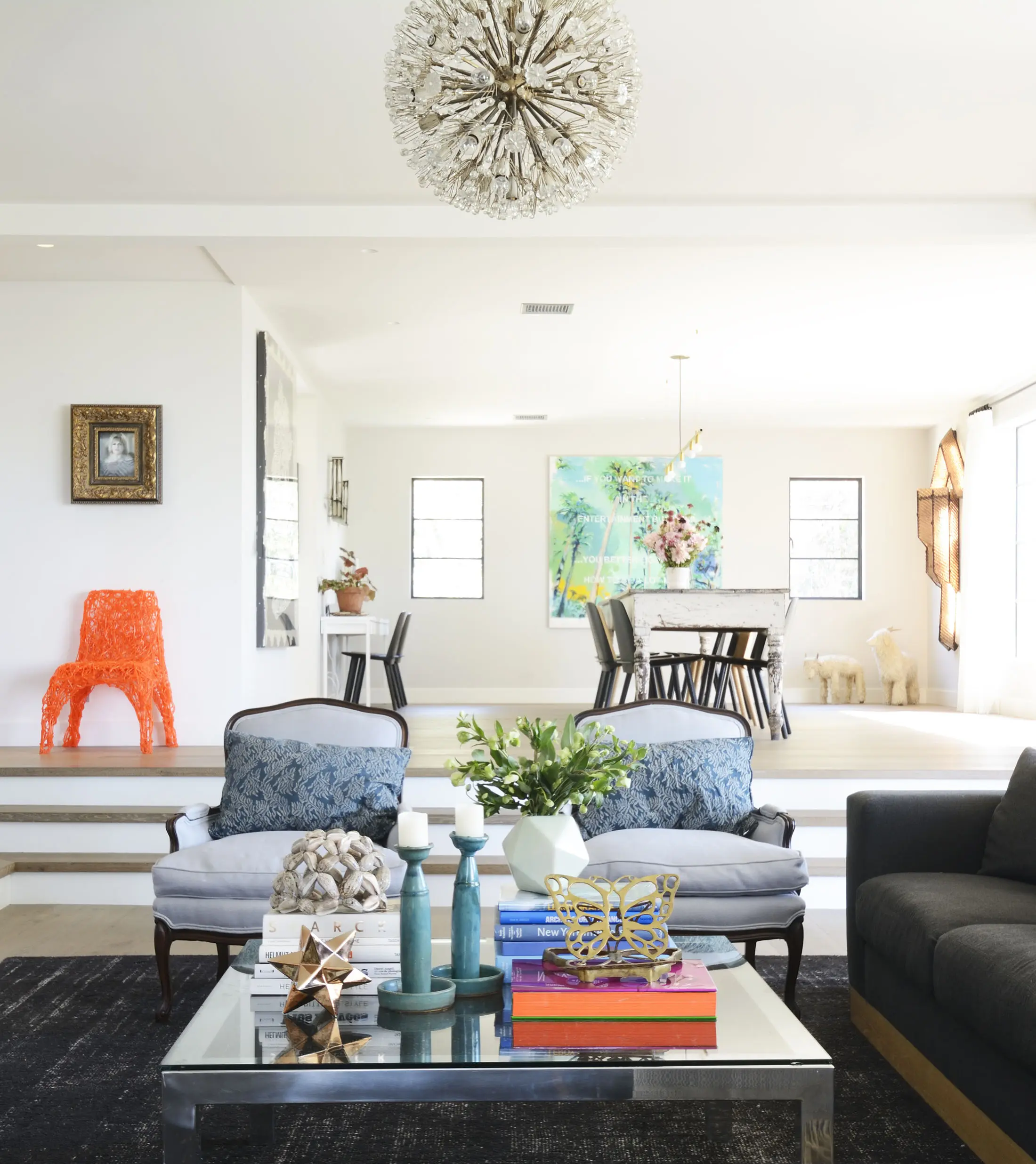 Spacious living room with stylish decor. Features a glass coffee table with books, candles, and flowers. Two blue cushioned chairs face a black sofa. Vibrant art and an orange chair are in the background. Above, a spherical chandelier hangs from the ceiling, illuminating the room.