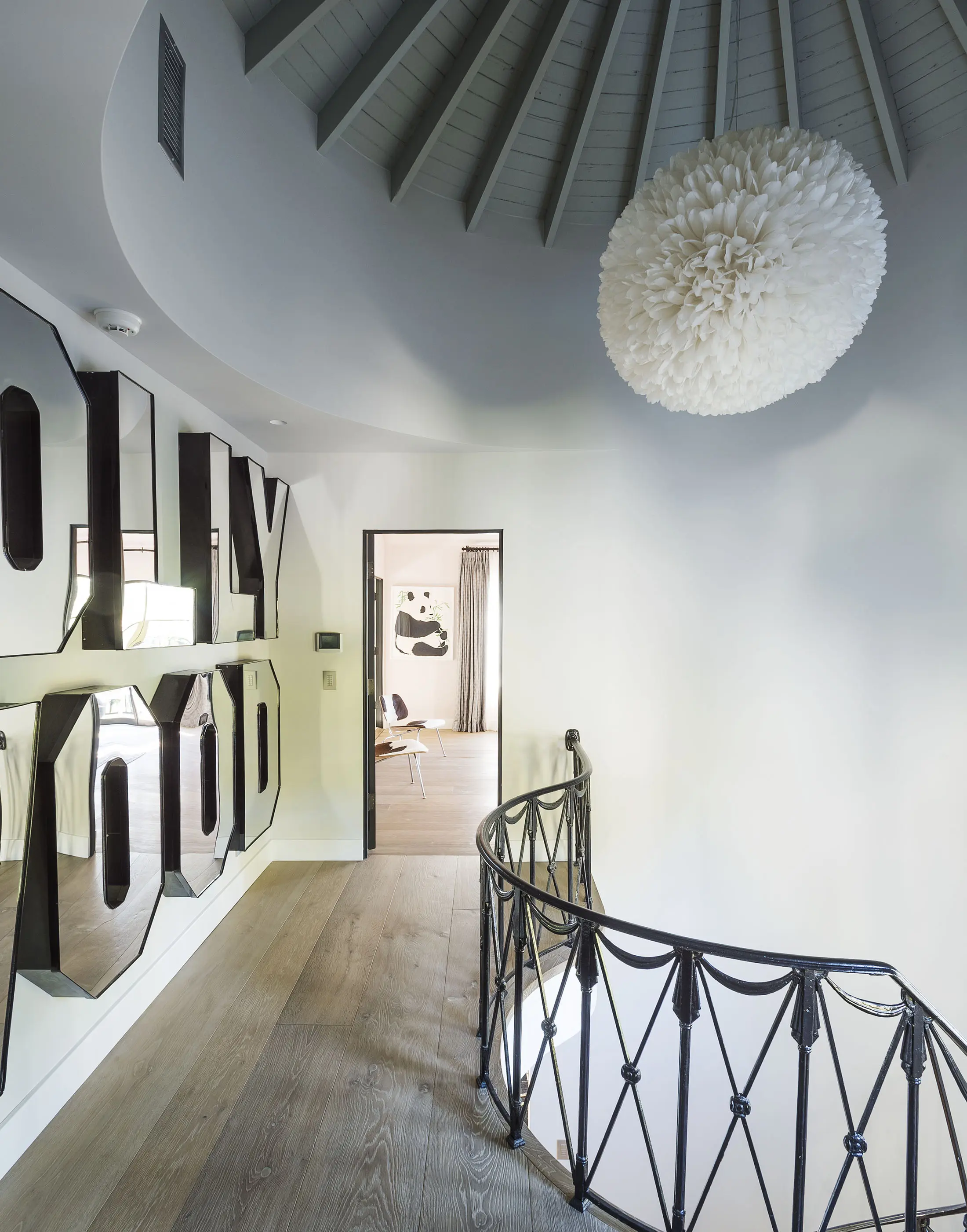 Spiral staircase with black railing leading to a room with a large artistic wall piece spelling HOLLYWOOD in mirrored letters. The ceiling features a large, round, white chandelier. A doorway opens to a room with a cow-themed mural visible in the distance.