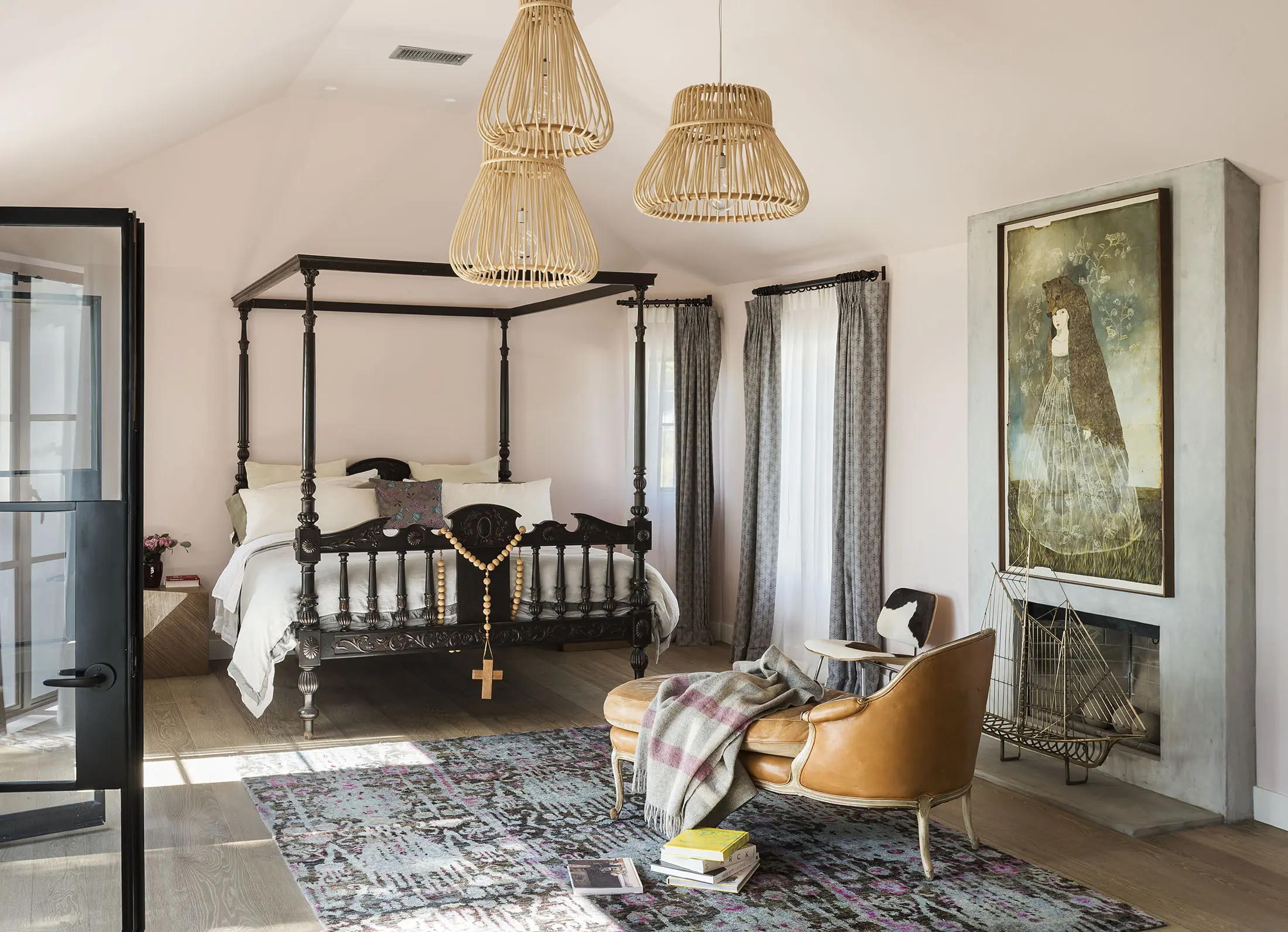 A bedroom features a dark wood canopy bed with white linens. A brown leather chair and ottoman face a fireplace with a patterned rug below. Above, three wicker pendant lights hang from the ceiling. A large framed artwork is on the right wall, and there are long gray curtains on the windows.