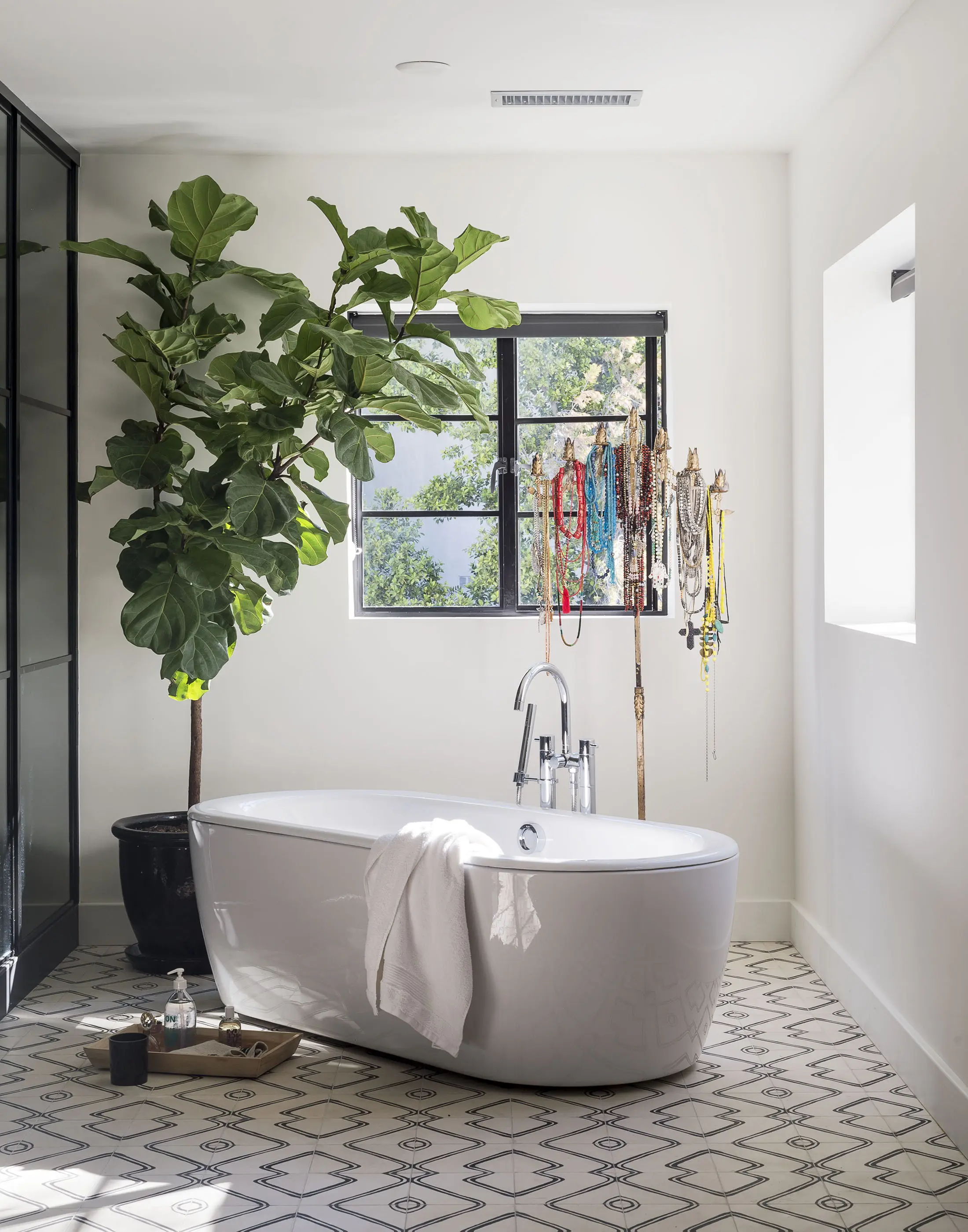 A modern bathroom with a white freestanding bathtub and a towel draped over the side. To the left, a large potted plant stands beside a window adorned with hanging jewelry. The tiled floor features a geometric pattern, and natural light streams through the window, creating a serene atmosphere.