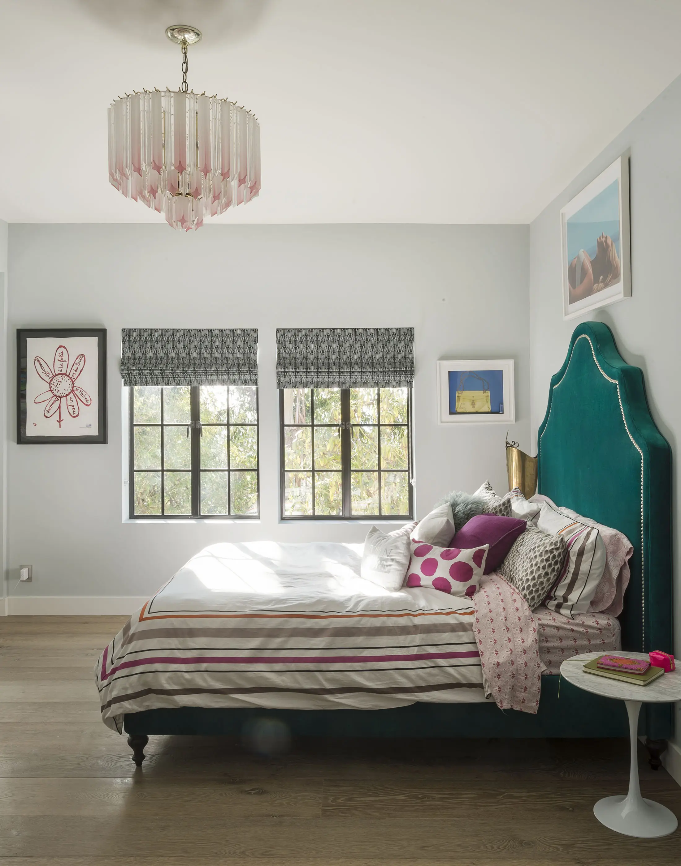 Bedroom with a teal upholstered bed adorned with colorful pillows and a striped duvet. Above, a chandelier featuring pink crystals hangs from the ceiling. The room includes artwork, two windows with patterned blinds, light gray walls, and a round white side table.