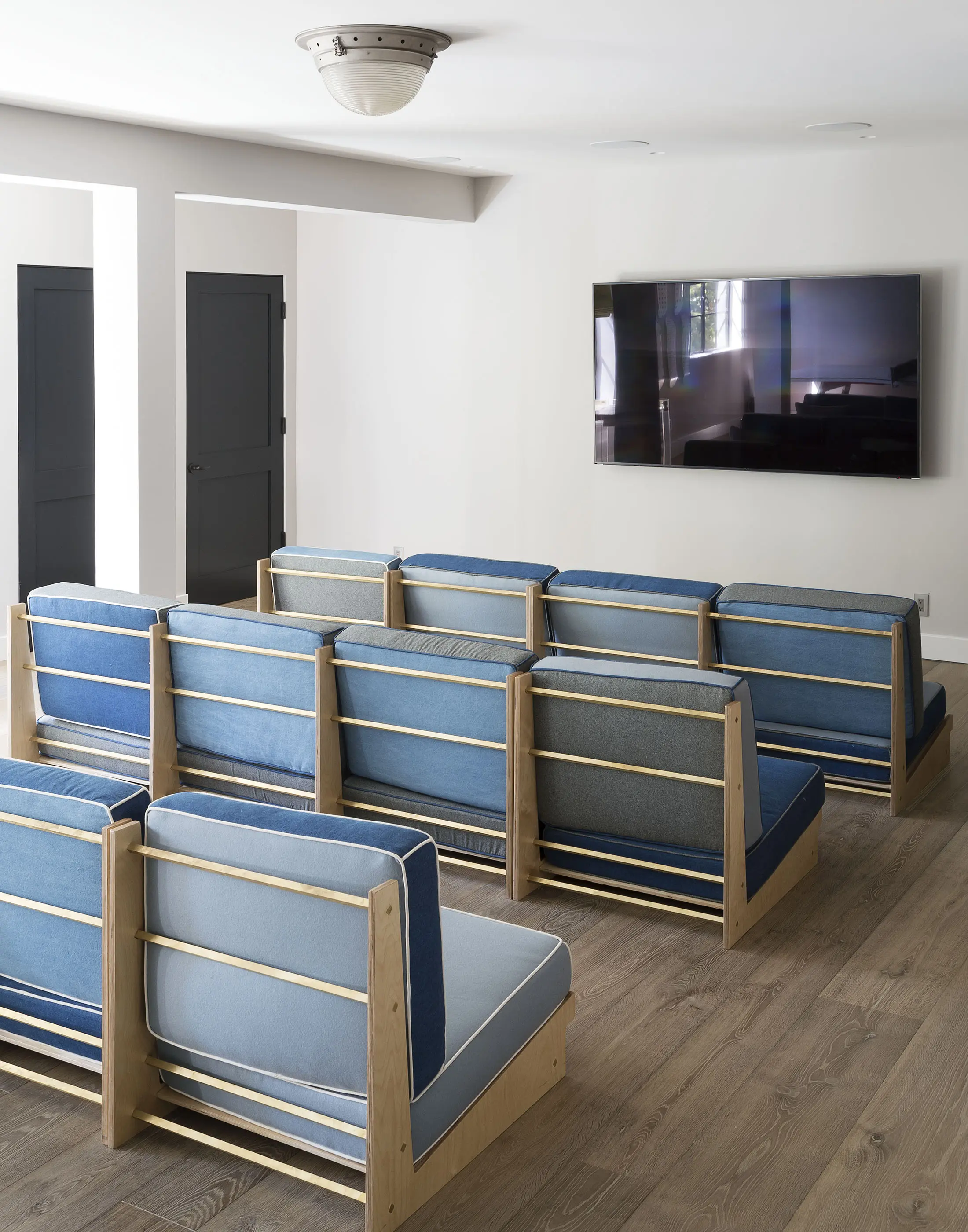 A modern home theater with three rows of blue cushioned seating. The seats have wooden frames with horizontal slats. A large, flat-screen TV is mounted on the light gray wall. Two black doors are visible on the left, and the floor is made of light wood. A ceiling light fixture is centered above.