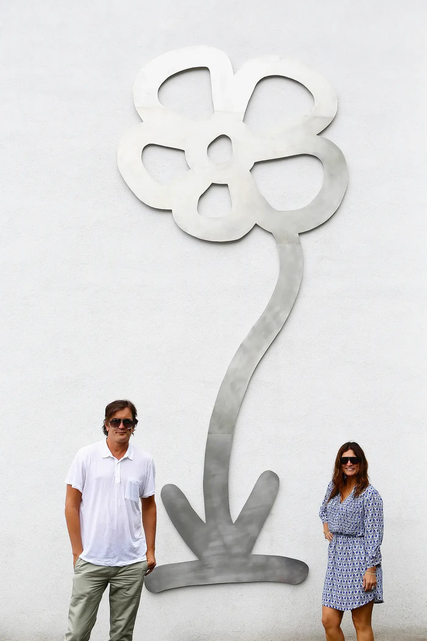 A man in a white shirt and sunglasses, and a woman in a blue patterned dress and sunglasses stand in front of a large metallic flower sculpture on a white wall. The sculpture features a simple, abstract design with a curvy stem and five petals.