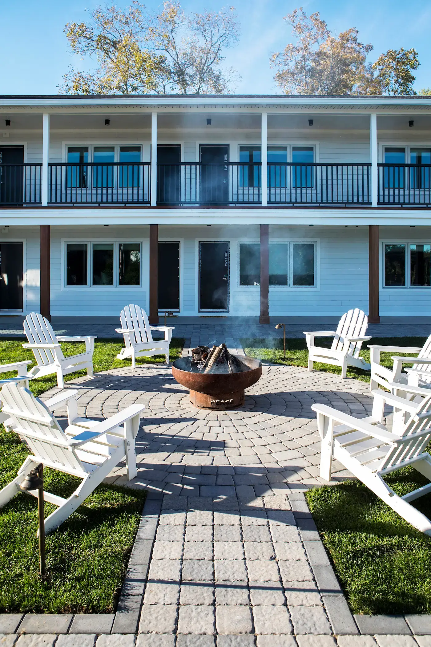 Outdoor fire pit area with Adirondack chairs arranged in a circle on a stone patio. Smoke rises gently from a metal fire pit. The backdrop features a two-story building with white siding and dark railing balconies, set against leafy trees under a clear sky.