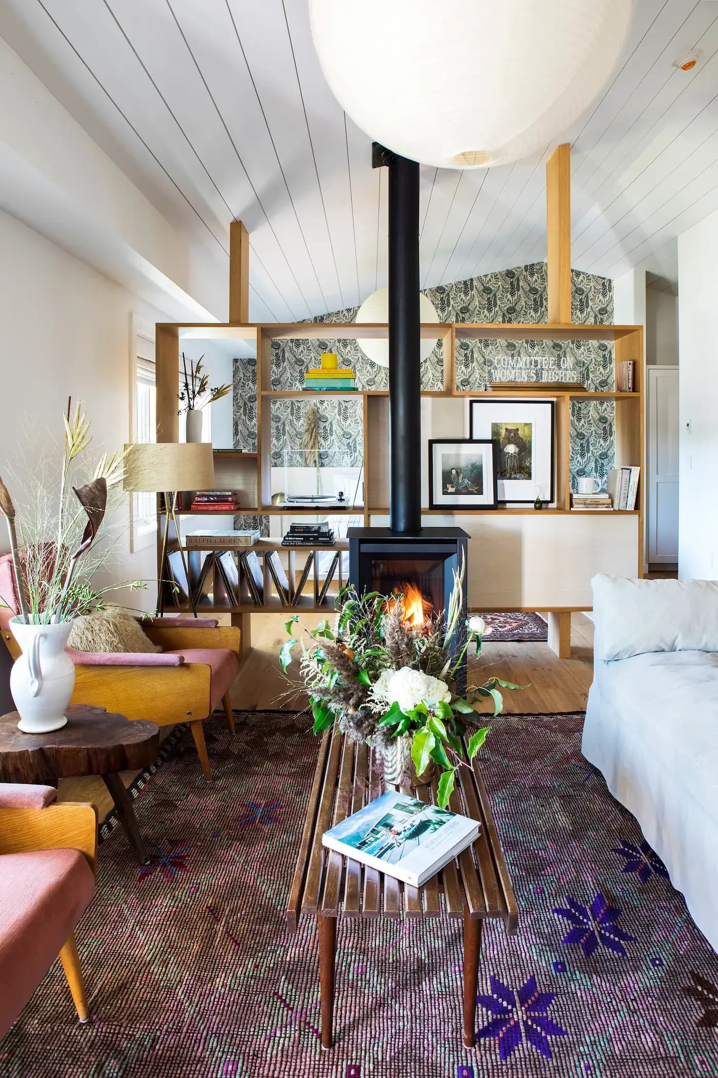 A cozy living room features a central wood-burning stove with a visible flame, surrounded by modern wooden furniture. Theres a rustic coffee table with plants, patterned rug, pink armchairs, and a white sofa. A bookshelf with books and framed art is in the background.