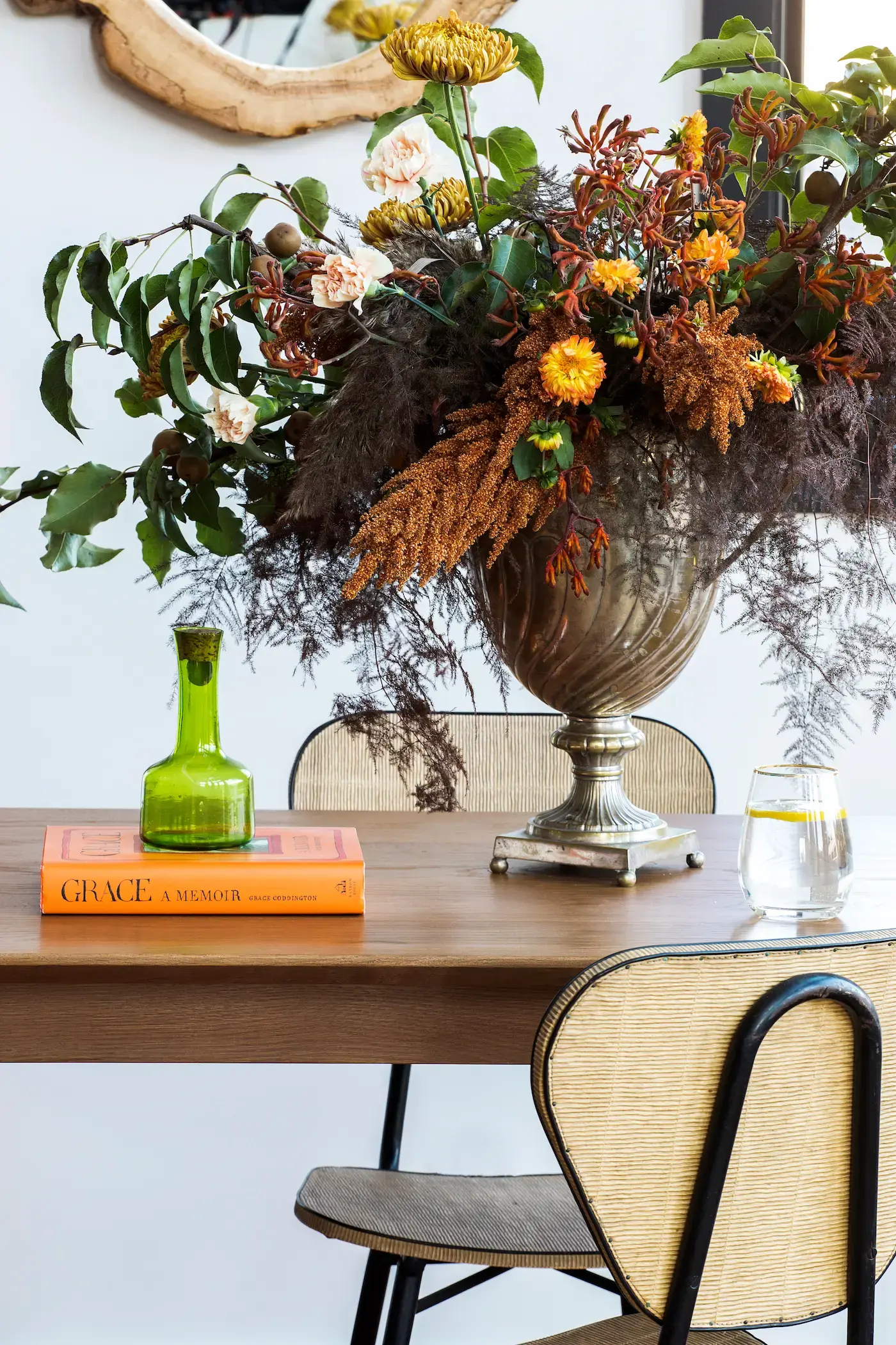 A large, ornate, silver vase on a wooden table holds an arrangement of yellow, orange, and pink flowers, with abundant foliage. Nearby, a green glass vase and an orange book titled Grace: A Memoir rest on the table. A glass of water is on the right, next to a wicker-backed chair.