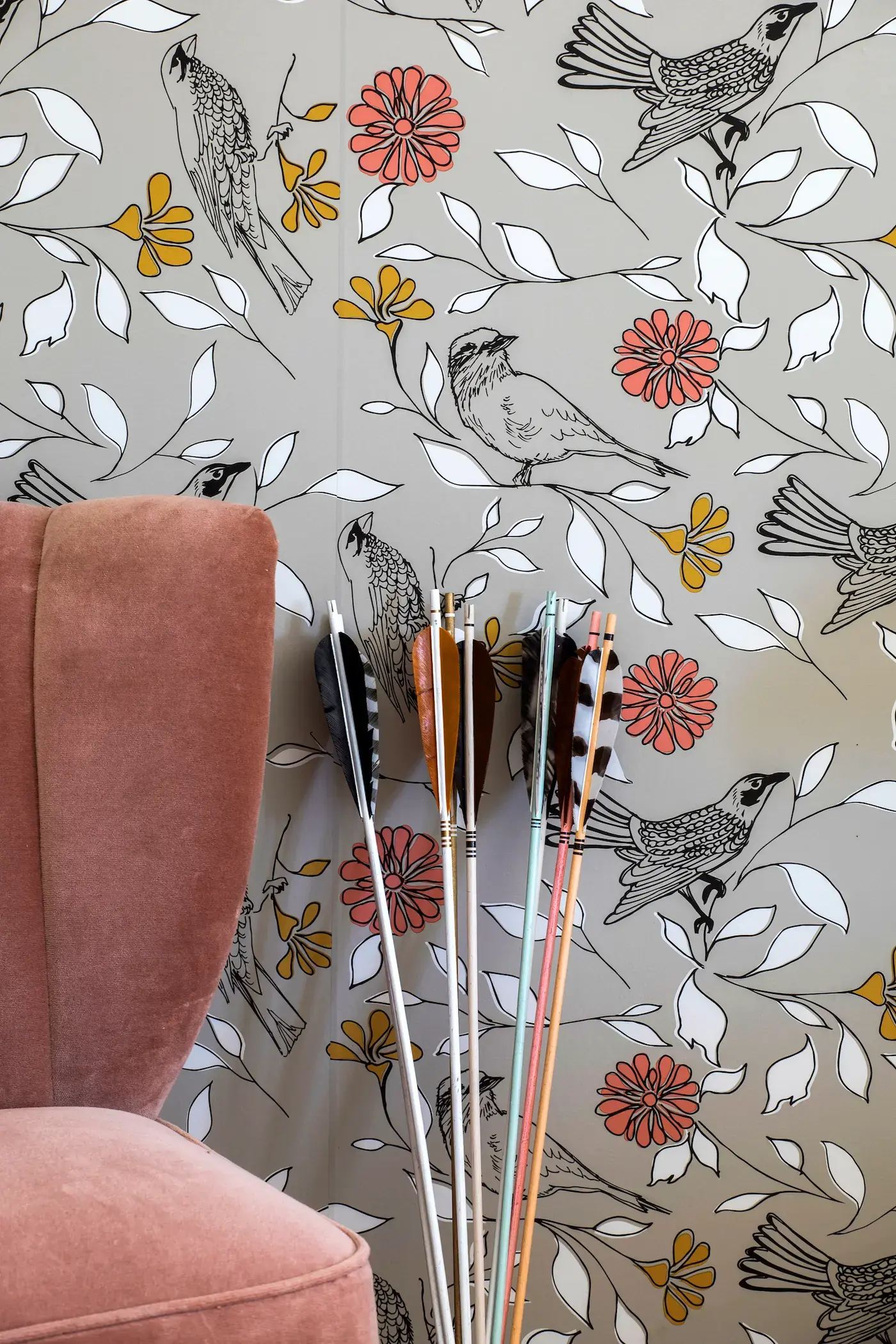 Close-up of a wall with bird and floral patterned wallpaper featuring red and yellow flowers. A velvet pink chair is partially visible on the left. Several arrows with various colored fletchings are grouped against the wall on the right.