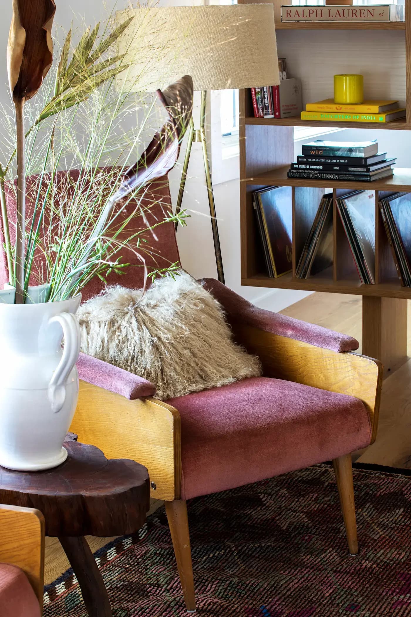 A cozy room with a vintage pink armchair featuring wooden armrests and a fluffy beige cushion. Nearby, a wooden shelf holds books and vinyl records. A white vase with tall grass sits on a rustic wooden table, and a floor lamp with a beige shade is in the background.