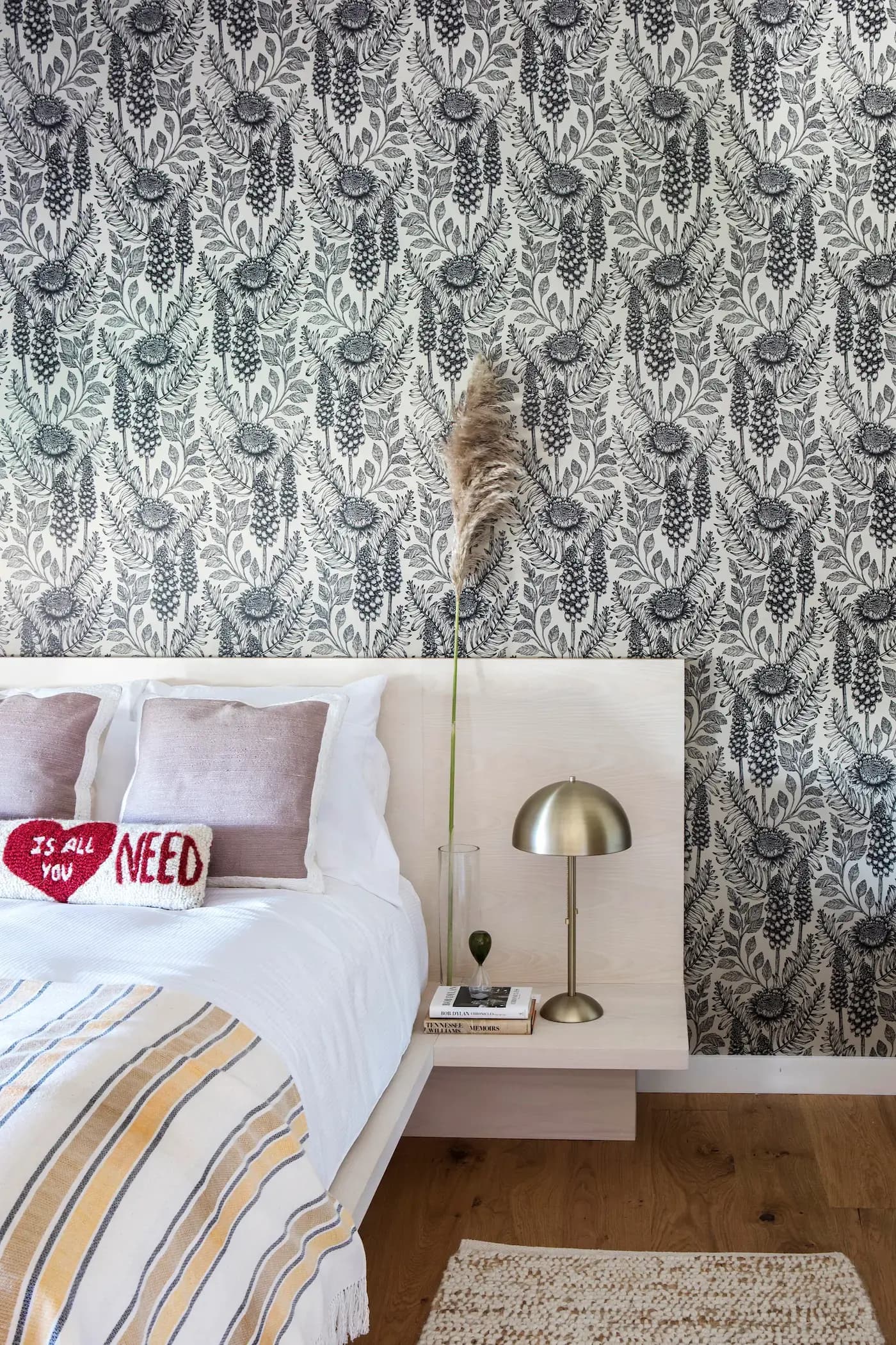Bedroom with a patterned black and white leafy wallpaper. A bed with white sheets and pillows, including one heart-shaped pillow reading ITS ALL YOU NEED. A bedside table holds a modern silver lamp and a tall vase with pampas grass. A textured rug lies on the wooden floor.