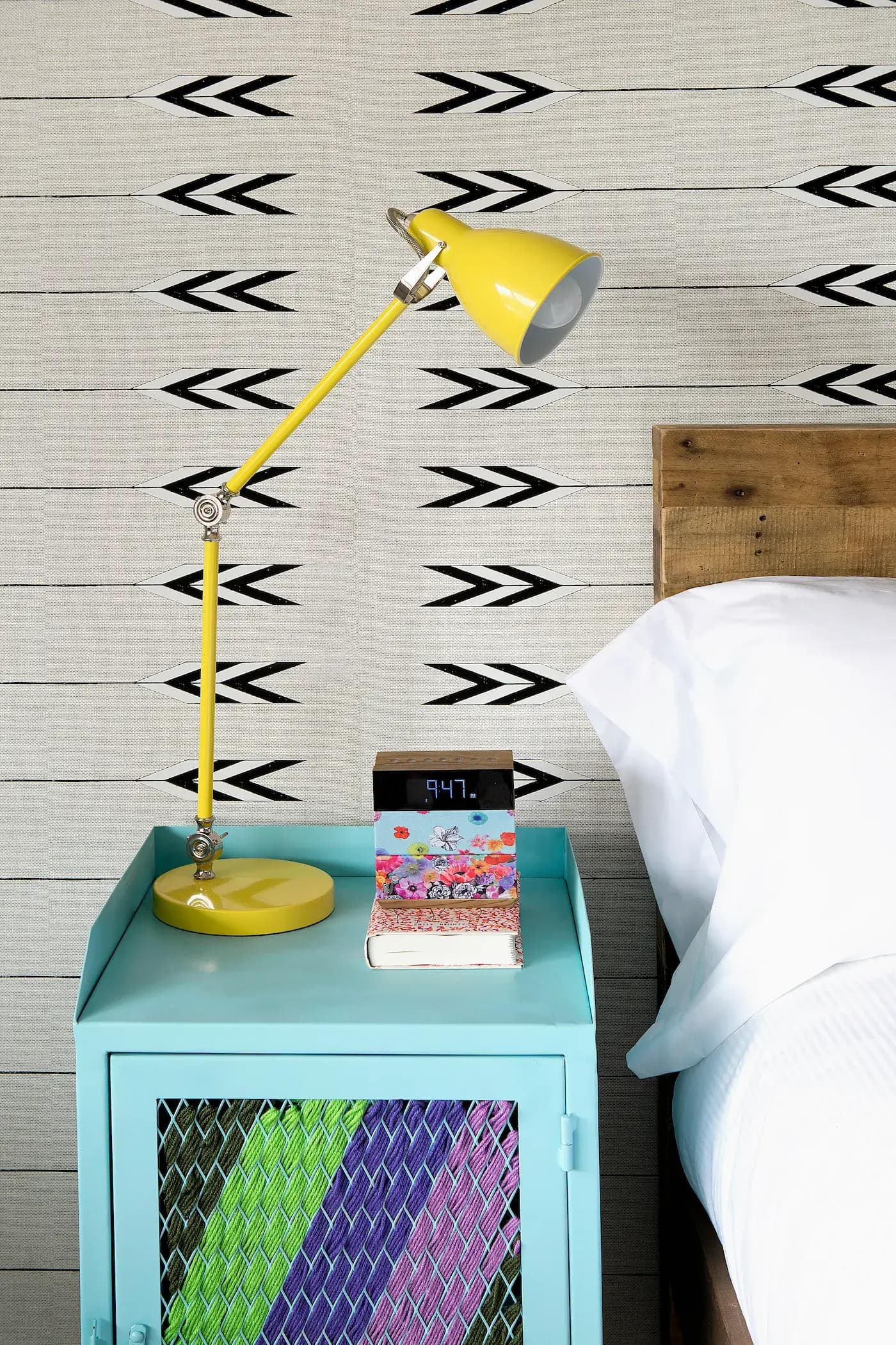 Yellow desk lamp on turquoise nightstand with colorful woven front. Alarm clock reads 9:47, resting on a floral box. Wooden headboard and white pillow in view. Wallpaper features black arrow pattern on beige background.