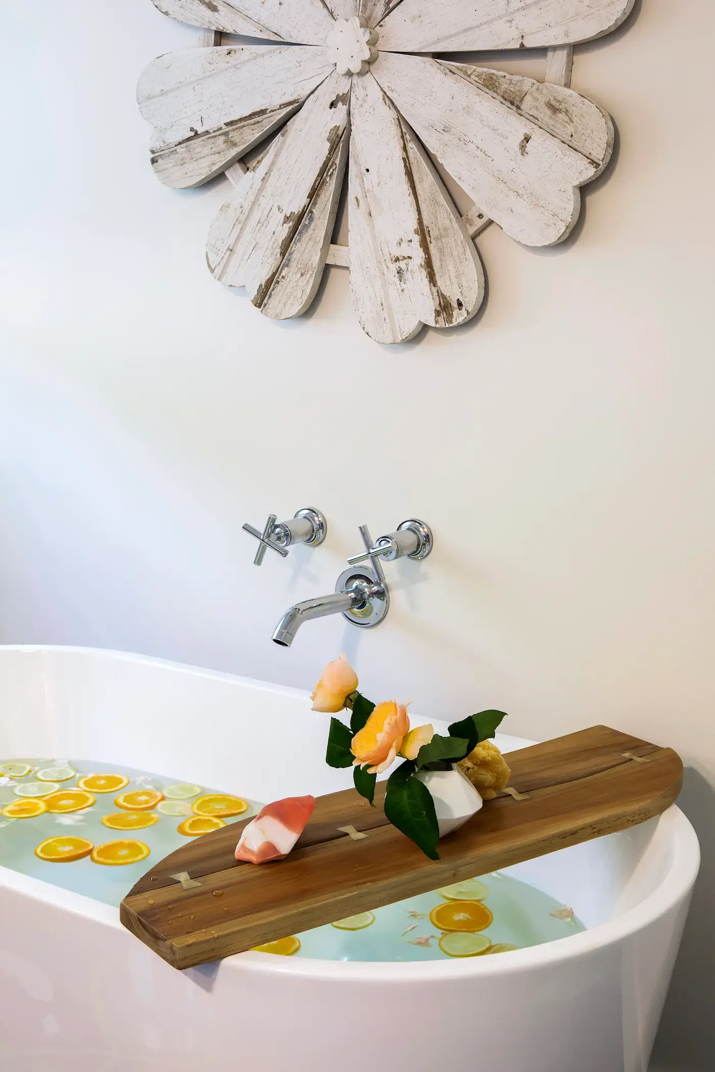 A white bathtub filled with water containing orange slices beneath a mounted wooden board holding orange and pink roses, with a tap and cross-shaped faucet handles above. A rustic, white wooden flower decor hangs on the wall above the setup.