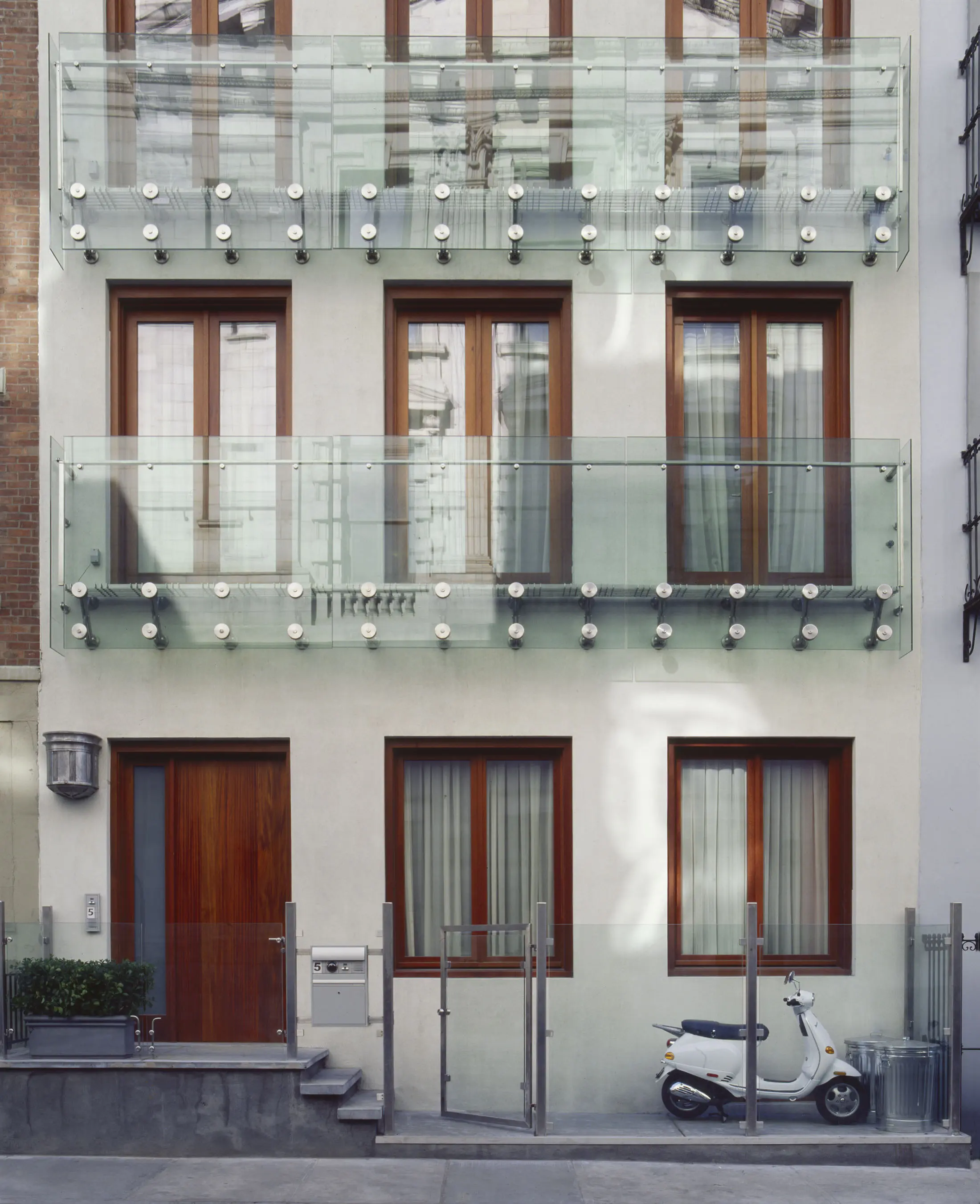 Modern building facade with three levels, each featuring large windows and glass balconies supported by metal. A small white scooter is parked at the ground level next to metal railings and stairs. The buildings exterior is light-colored with wood accents and urban surroundings.