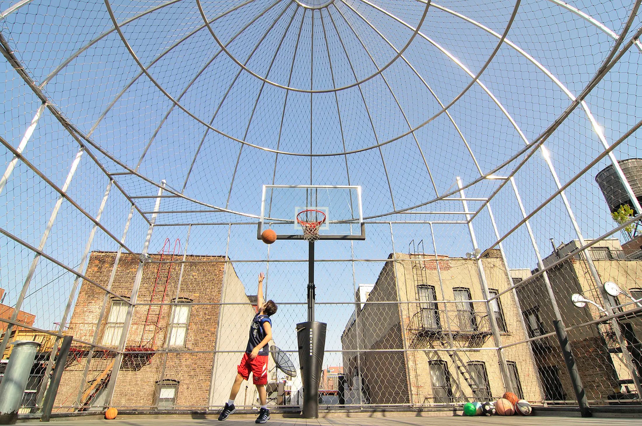 A person in a blue shirt and red shorts jumps to shoot a basketball on an outdoor court. The court is enclosed with a high, dome-shaped wire fence. Several basketballs are scattered on the ground. Surrounding brick buildings are visible under a clear blue sky.
