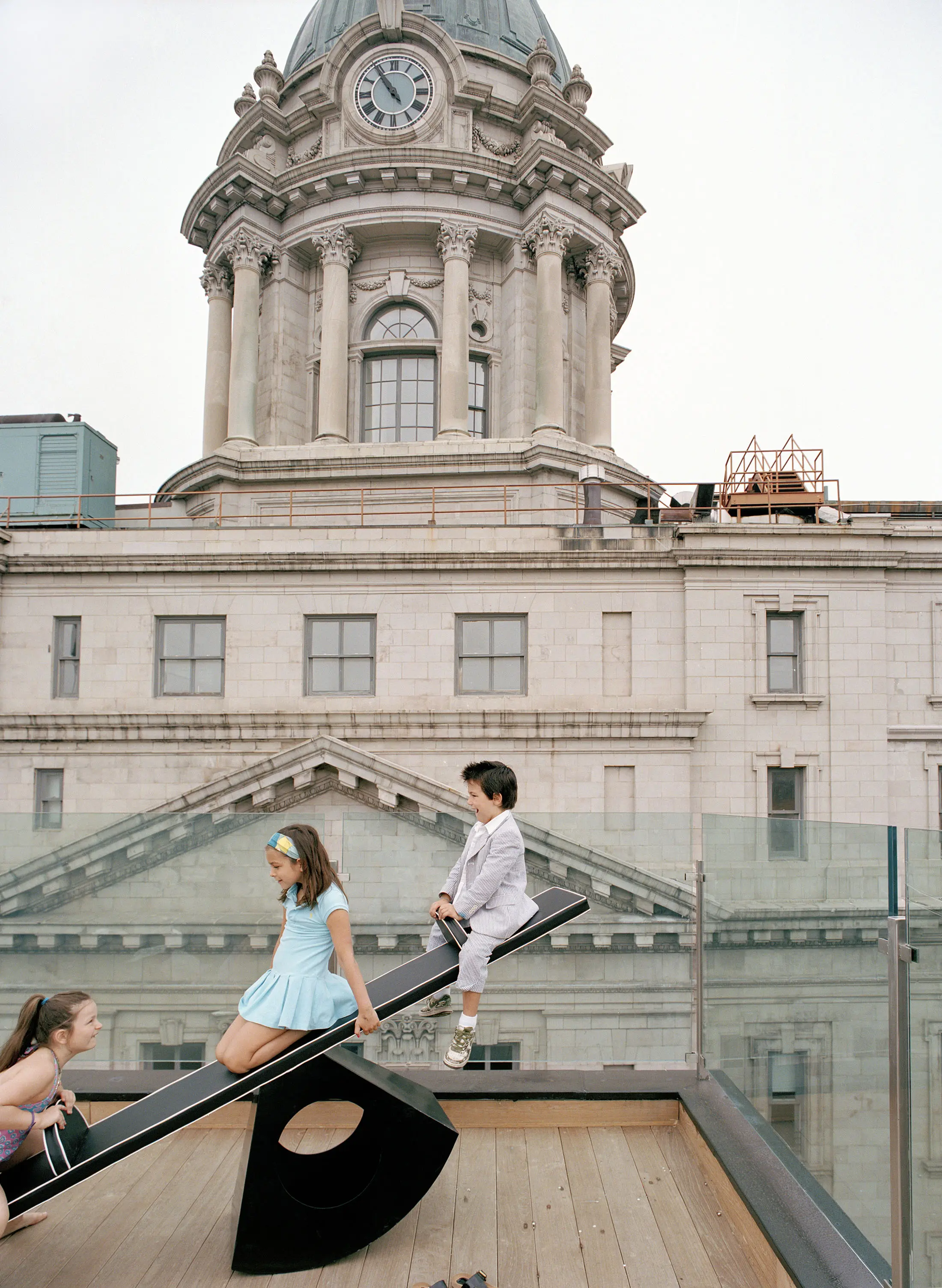 Three children play on a seesaw on a rooftop with a historic building in the background. The child on the left is in a purple outfit, the center child wears a blue dress, and the one on the right is in a gray suit. The building has a clock tower and classical architecture.