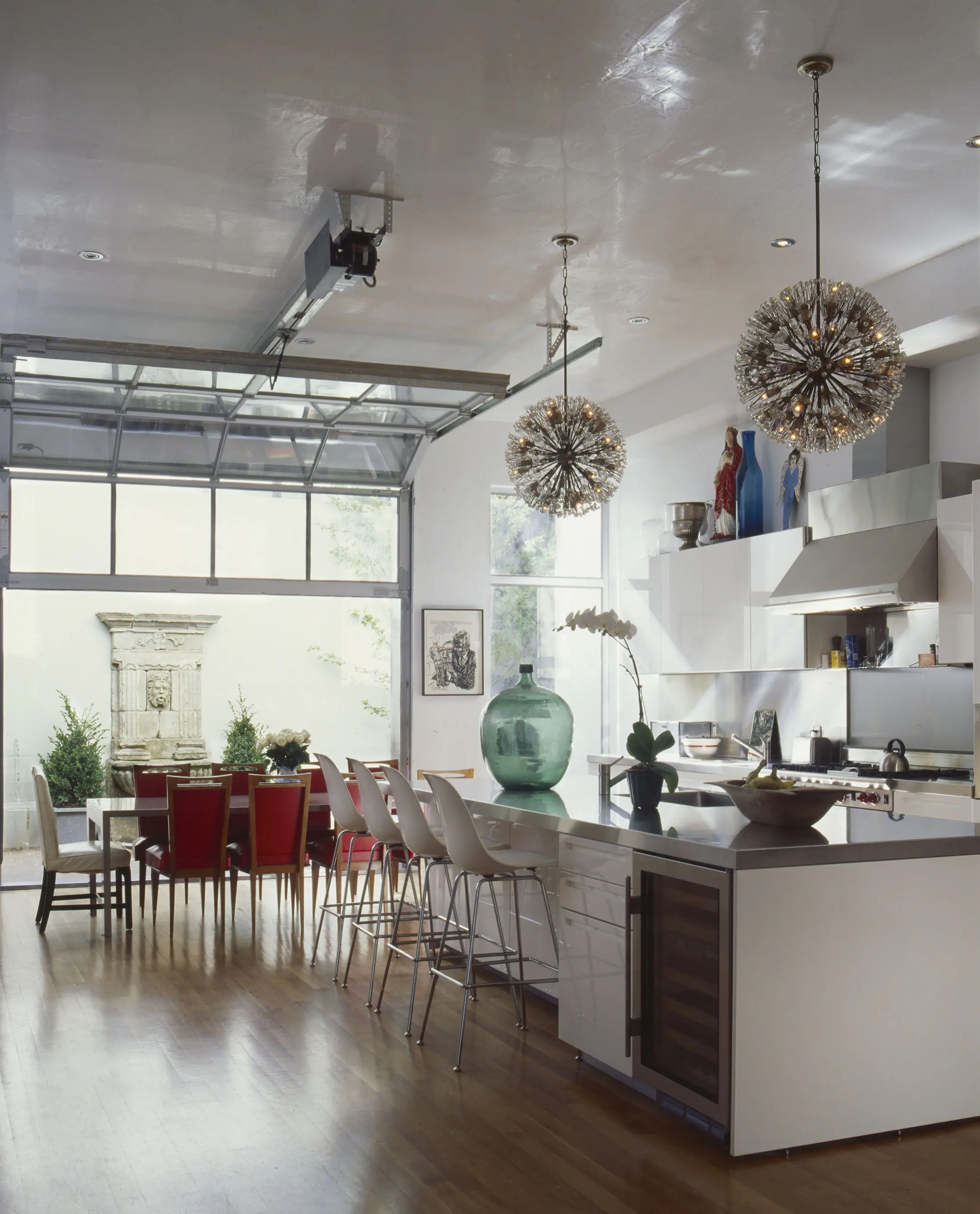 Modern kitchen and dining area with an open glass garage door. White cabinetry and a marble island with bar stools occupy the kitchen. Two spiky pendant lights hang from the ceiling. The dining table features red chairs. Large glass vase centerpiece on the island.