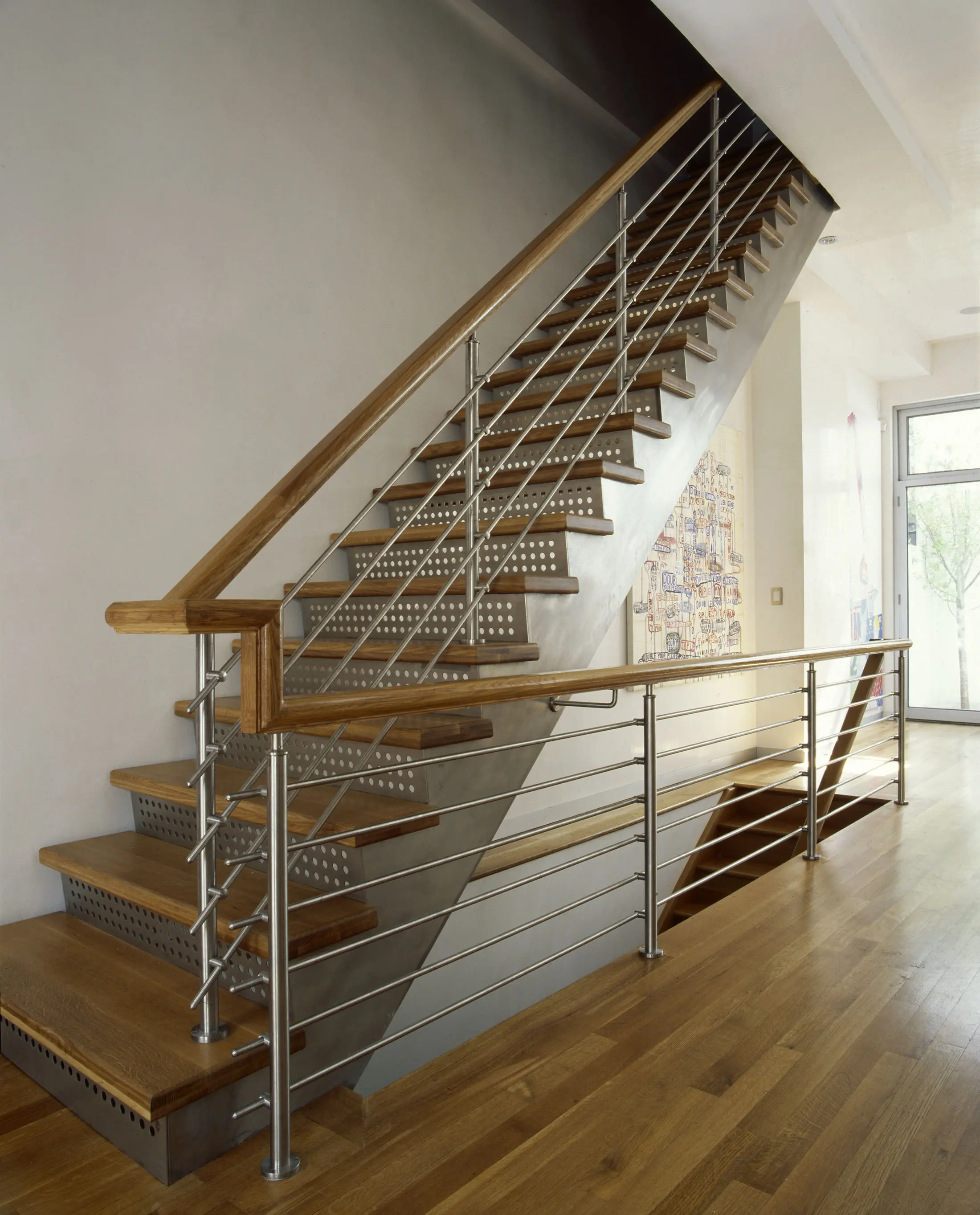 Modern wooden staircase with sleek metal railings against a gray wall. The stairs ascend to the upper floor, with natural light streaming in from nearby windows. The wooden floor and steps complement the minimalist design of the setting.