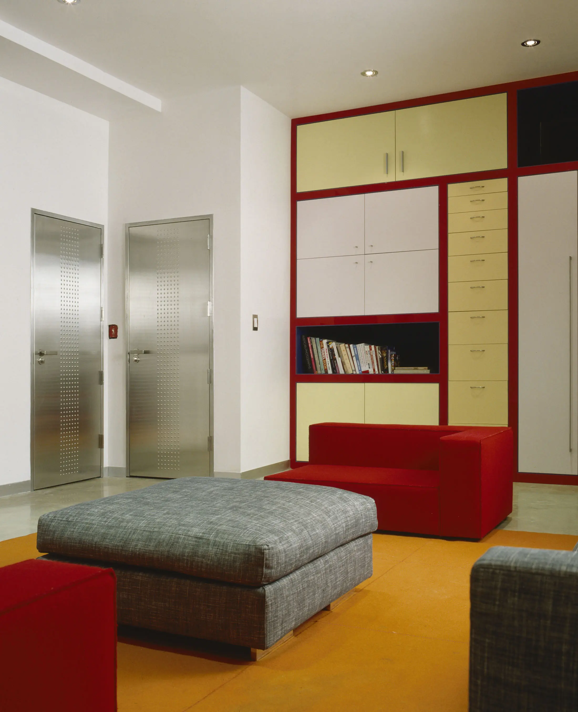 A modern room with red and gray furniture. There is a red chair and a gray ottoman on an orange carpet. The back wall features a large shelving unit with red trim, yellow drawers, and books. Two metallic doors are on the left, and the ceiling has recessed lighting.