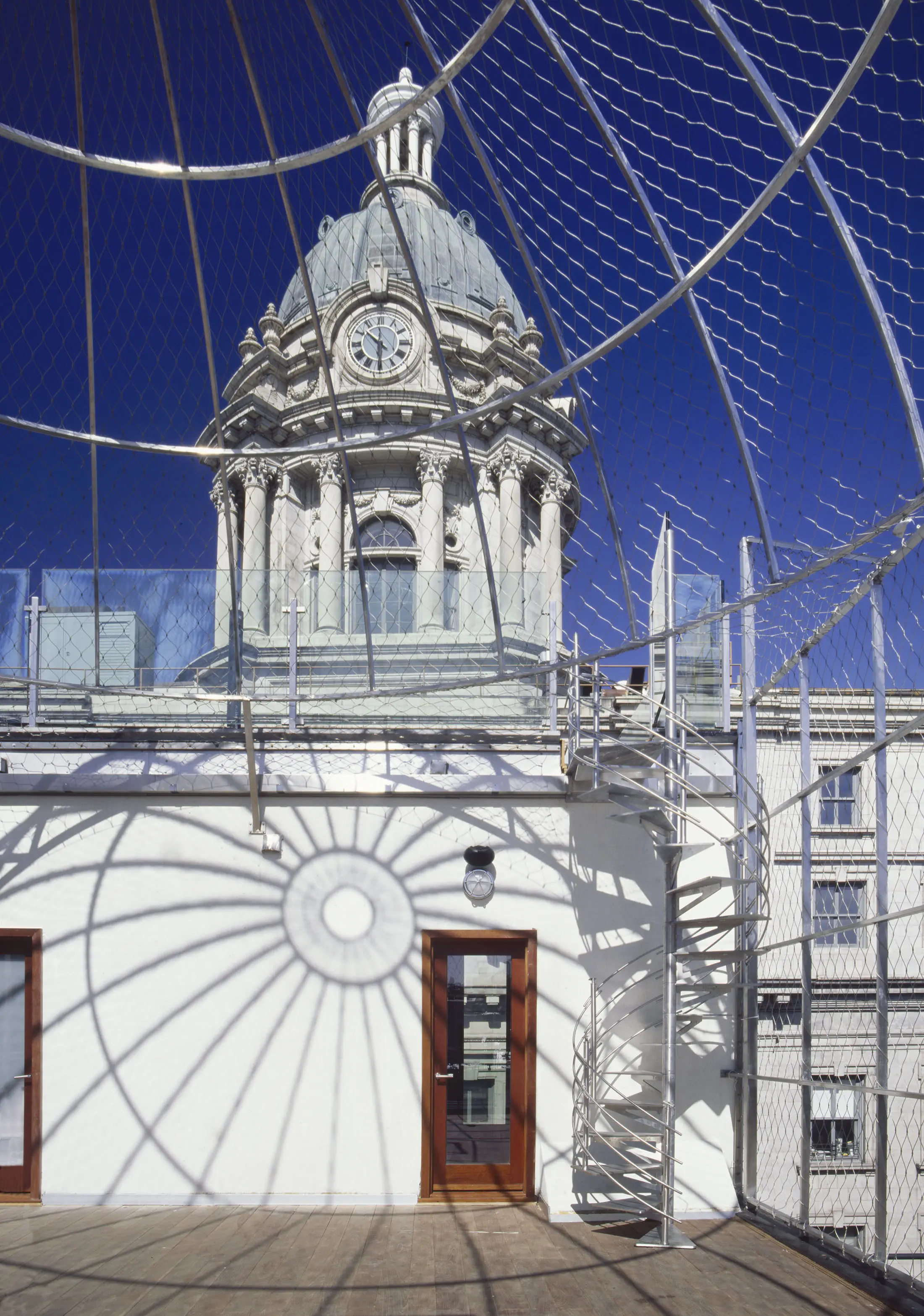 A rooftop with a metal mesh dome casting intricate shadows on the floor. A wooden door and light fixture are on a white wall. In the background, an ornate clock tower with columns and a dome is visible against a deep blue sky. A spiral staircase leads up the structure.