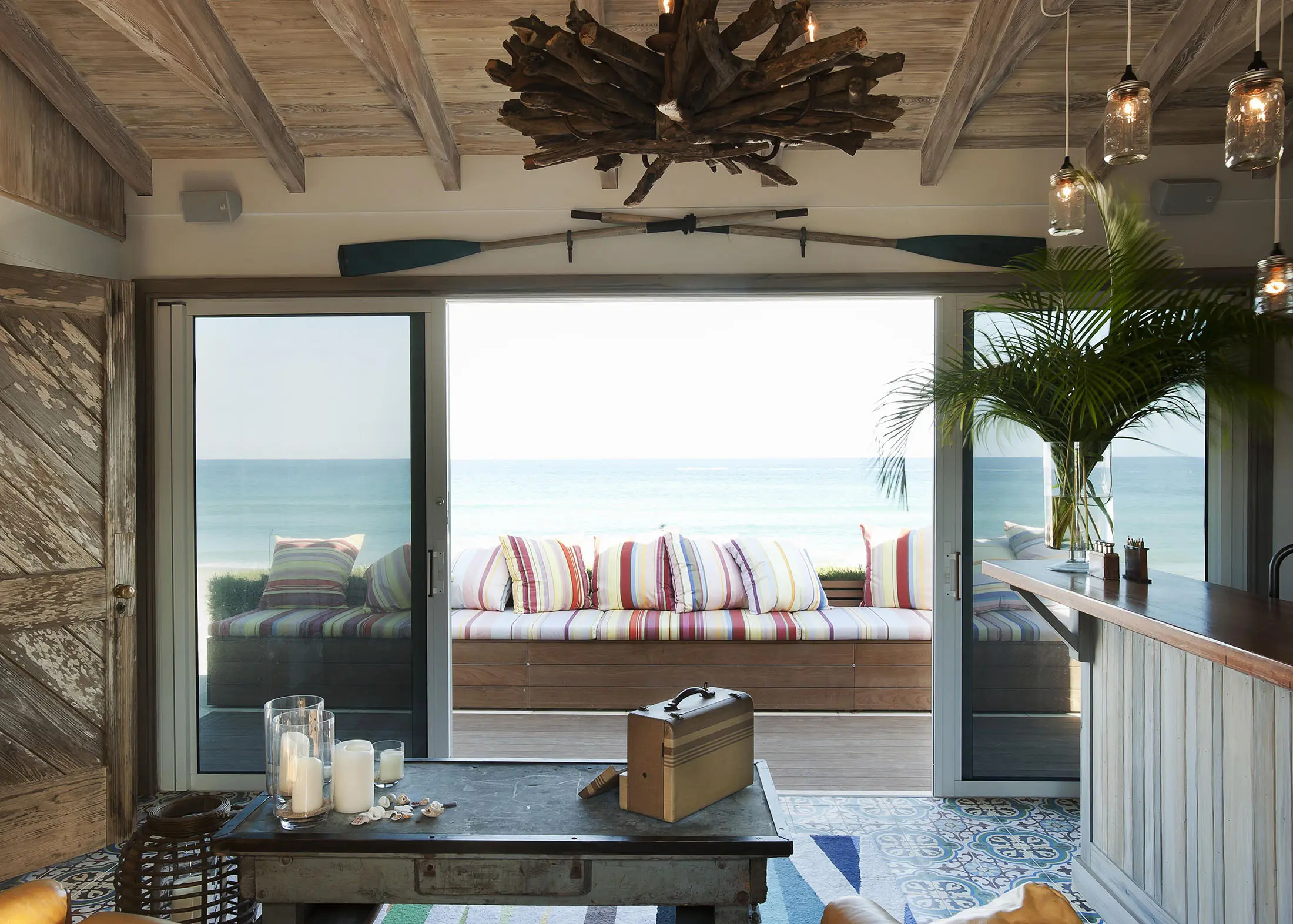 Beachfront room with a sliding glass door opening to a view of the ocean. Inside, theres a rustic wooden table with candles, a radio, and striped cushioned seating outside. A ceiling chandelier made of driftwood and glass lights hangs above. An oar decorates the wall.