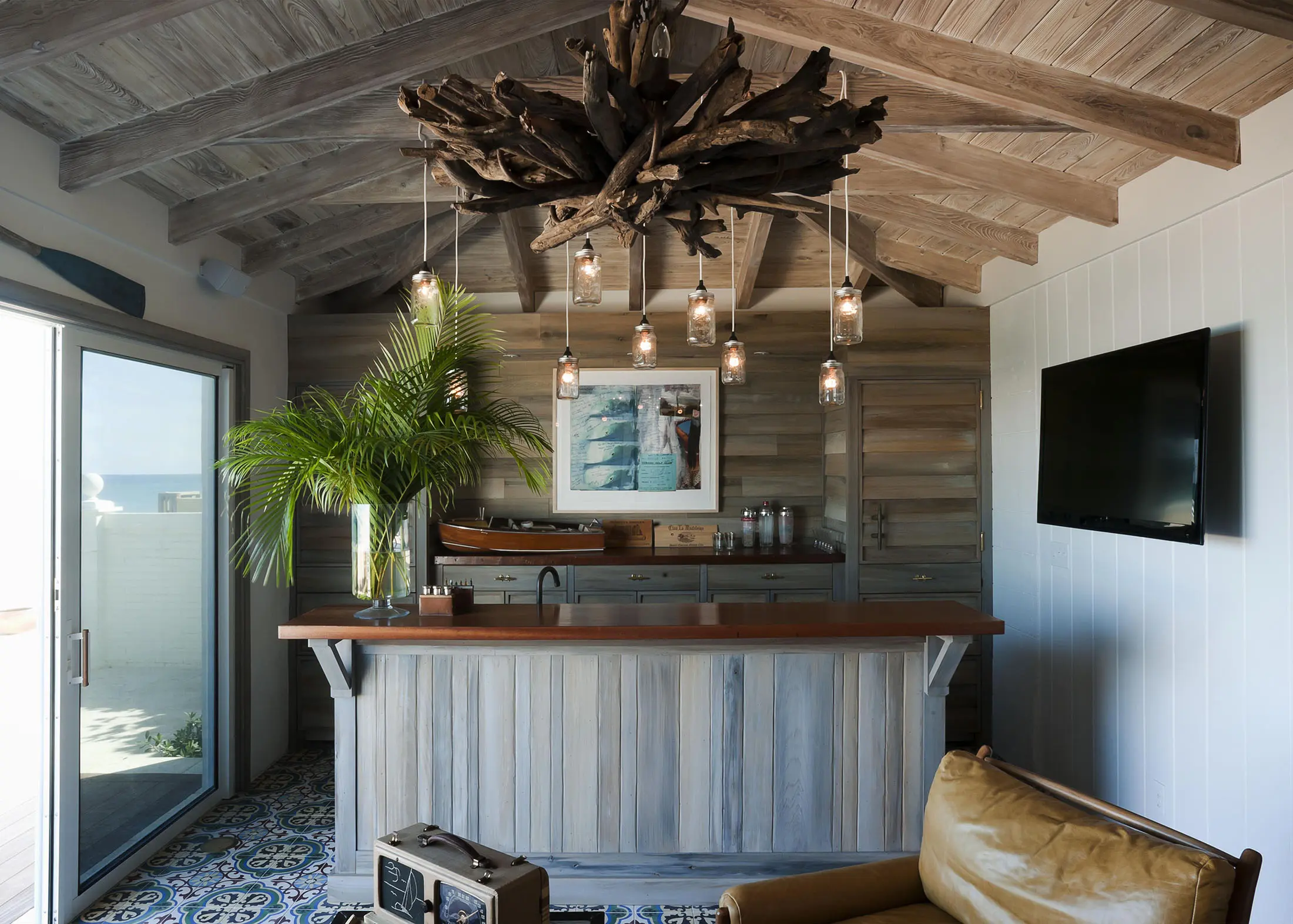 Cozy room with wooden beams and rustic decor. A bar with a wood countertop is the focal point, adorned with a driftwood chandelier. Theres a framed picture on the wall above the bar, a potted palm plant, a wall-mounted TV, and a leather chair on patterned tiles, plus sunlight from a glass door.
