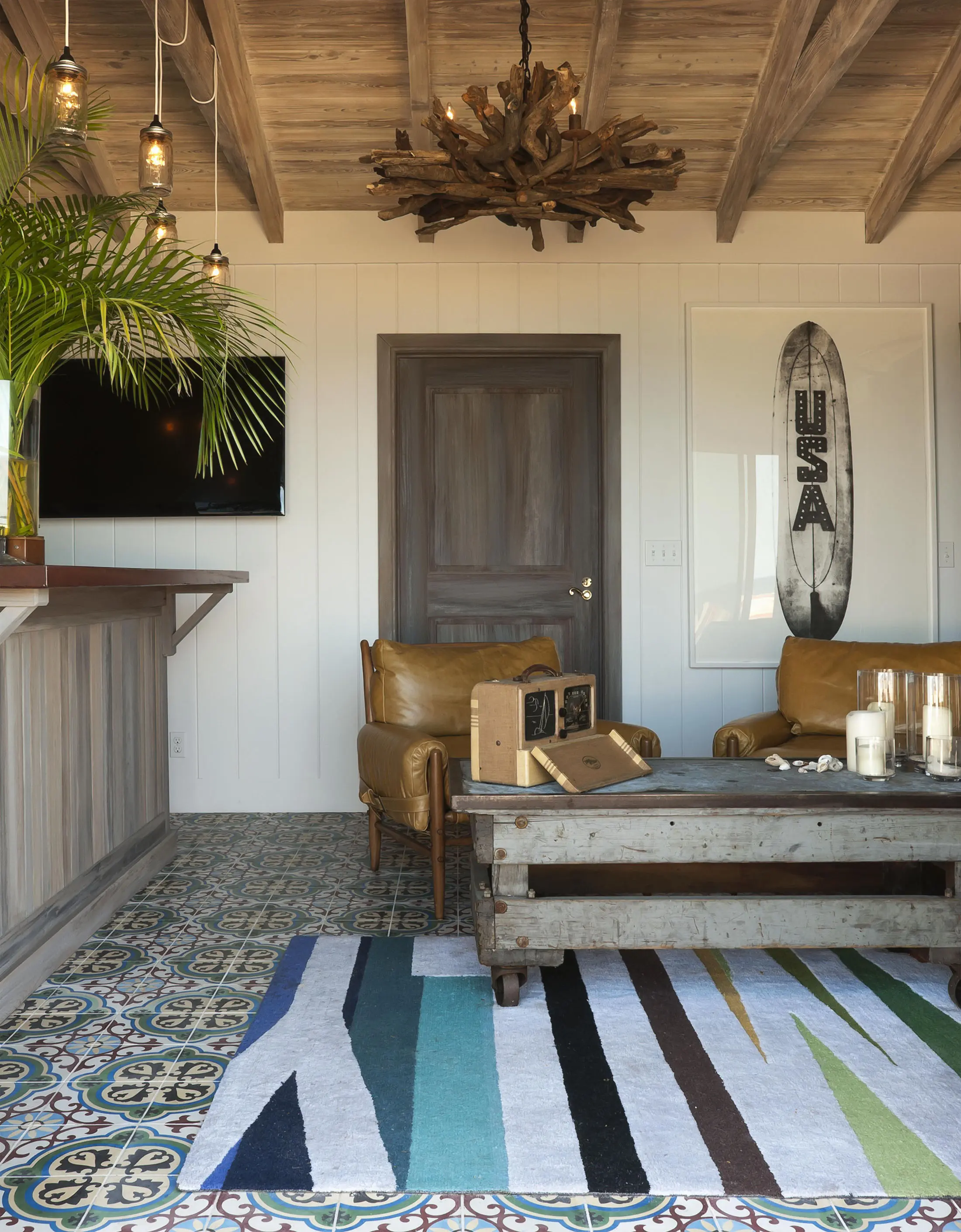 Rustic living room featuring leather chairs, a wooden coffee table, vintage surfboard art, and multi-colored rug on patterned tile floor. Driftwood chandelier hangs from wood-beamed ceiling. Wall-mounted TV and plants complete the cozy, eclectic decor.
