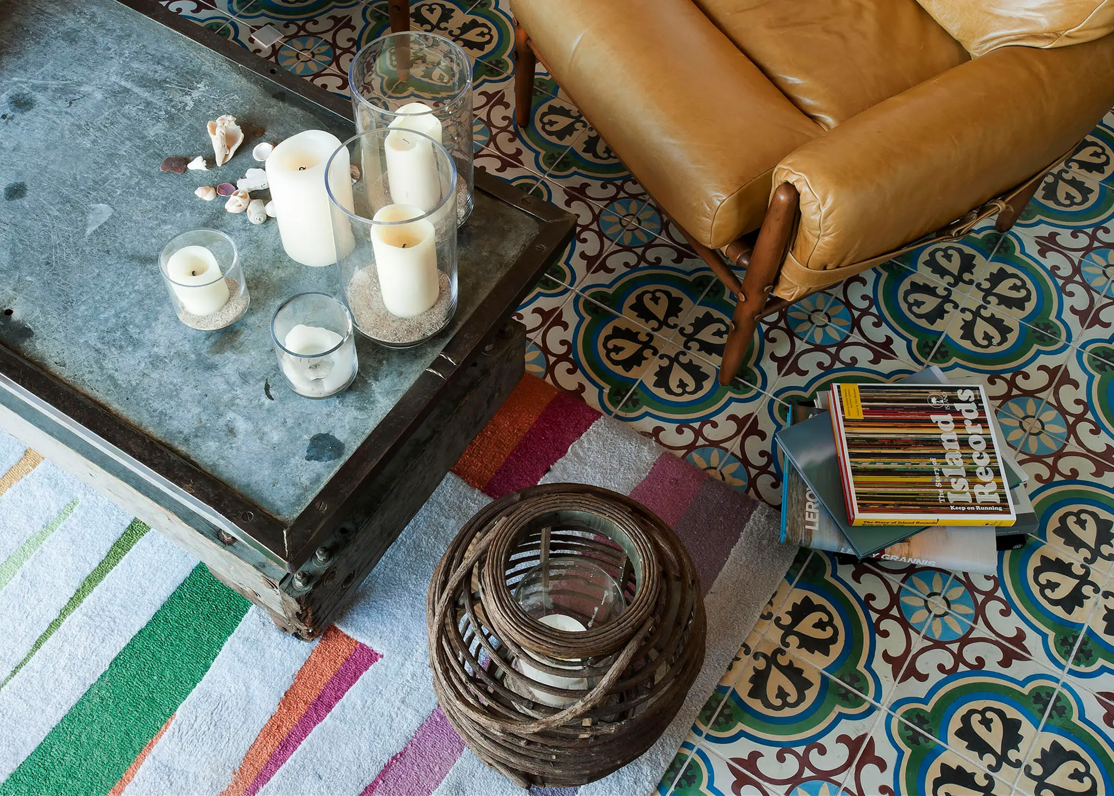 A living room scene with a metal coffee table holding white candles in glass holders and a few seashells. A tan leather chair sits on a patterned floor. A stack of magazines lies on the floor, next to a striped rug and a woven basket lantern. Colorful, intricate tiles cover the floor.
