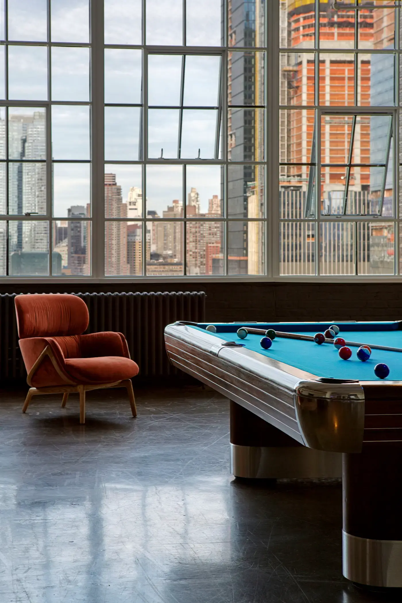 A dimly lit room with a pool table scattered with balls in the foreground. A rust-colored armchair sits nearby on a dark wooden floor. Large windows reveal an urban skyline outside, with tall buildings and cranes against a cloudy sky.
