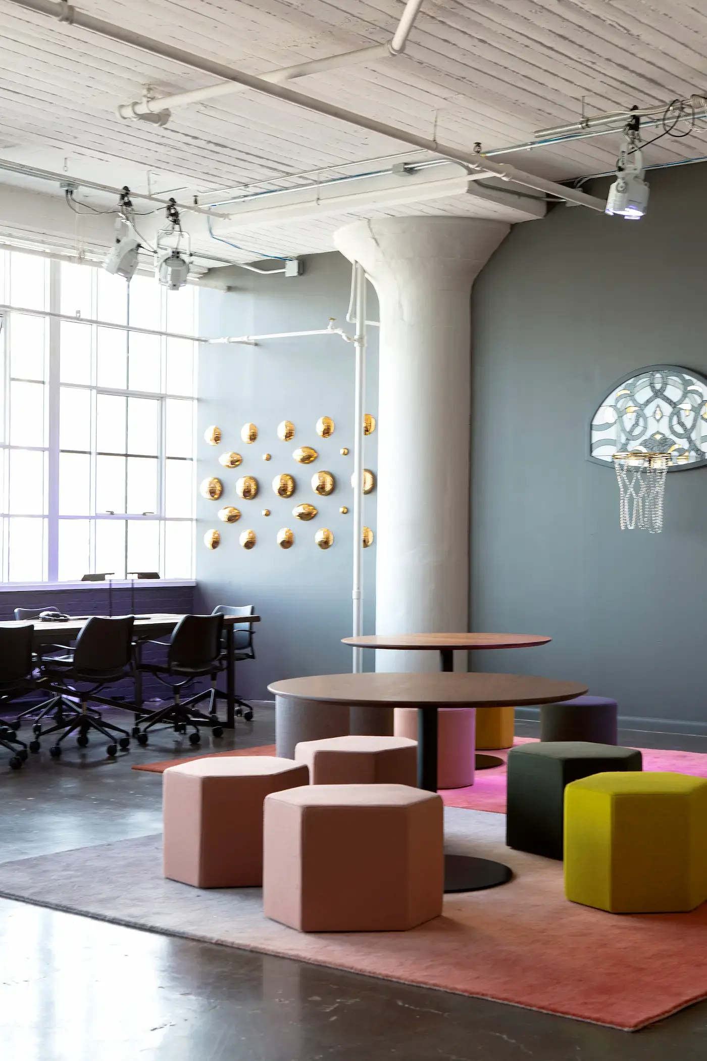 A contemporary room with a basketball hoop mounted on a gray wall. Theres a circular table surrounded by hexagonal stools in muted pink, green, and beige on a pink rug. A conference table with chairs is near large windows. Wall decor includes a cluster of circular gold accents.