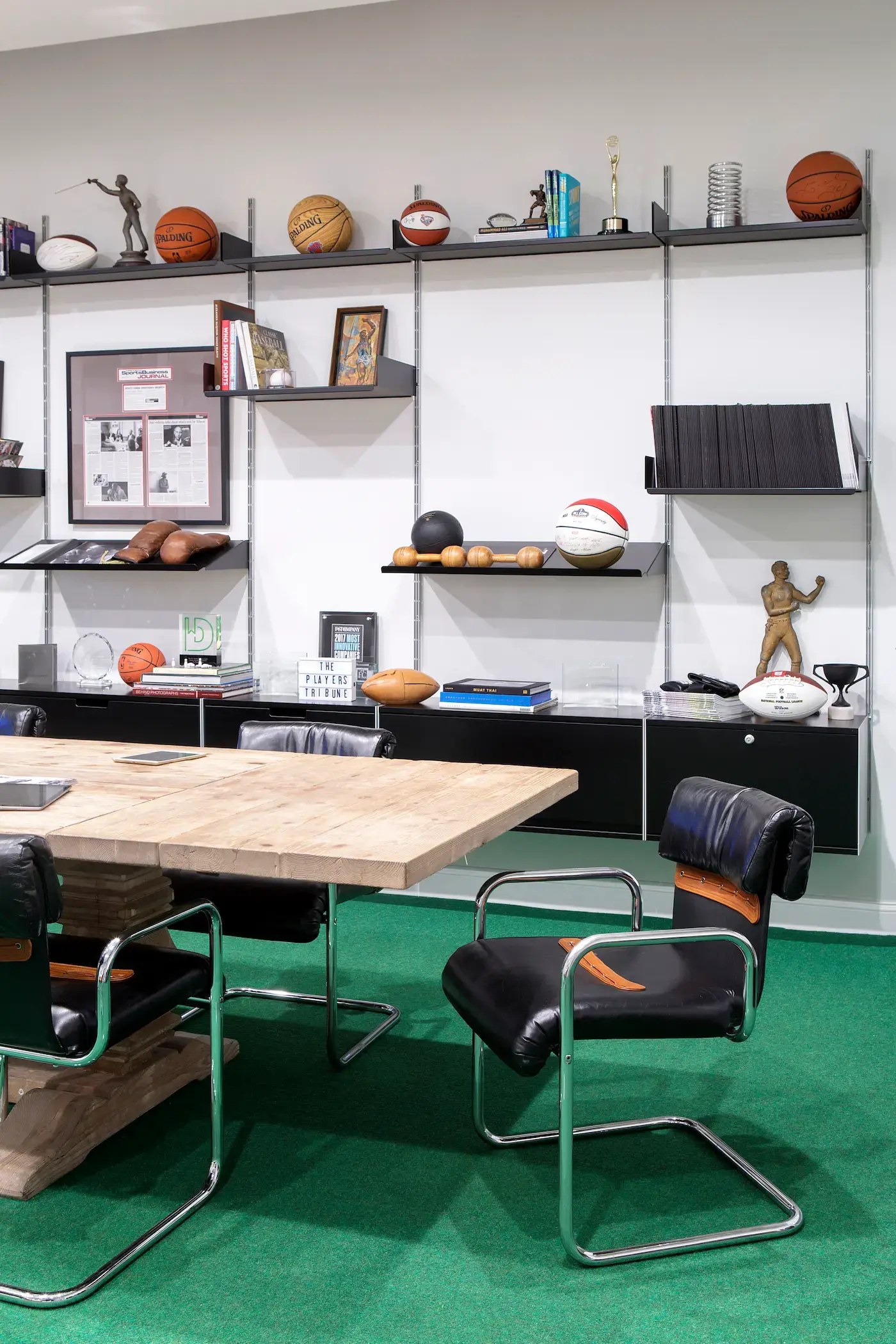 Office with a wooden table and black leather chairs on a green carpet. Shelves display basketballs, awards, and books. Framed pictures and memorabilia adorn the wall. A gold statuette and various sports items are showcased, creating a sporty, professional ambiance.