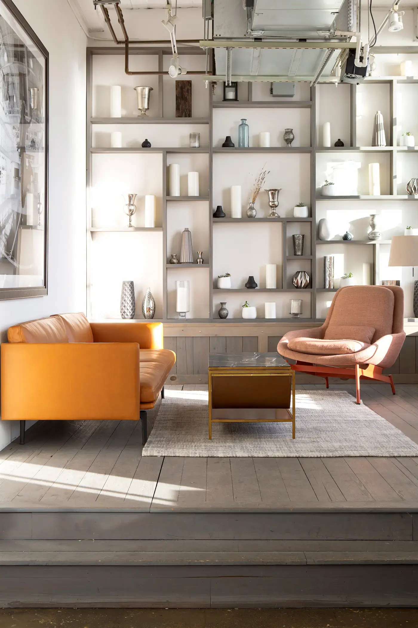 A modern living space with a brown leather sofa, a brown lounge chair, and a glass coffee table on a gray rug. The backdrop features a wall of shelves with various vases and decorative items. Exposed ceiling pipes and large artwork are visible. The room is well-lit with natural light.