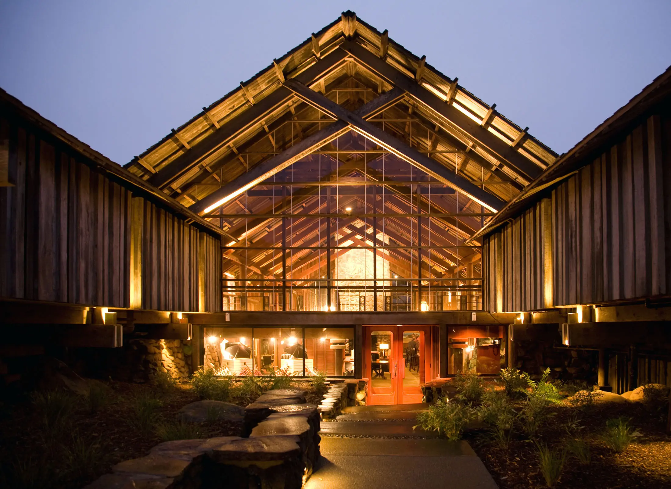 A lodge with a striking A-frame structure, showcases wooden and glass elements. Warm lighting illuminates the interior and exterior, highlighting the entrance and landscaped path. The evening sky provides a serene backdrop, complementing the rustic yet modern design.