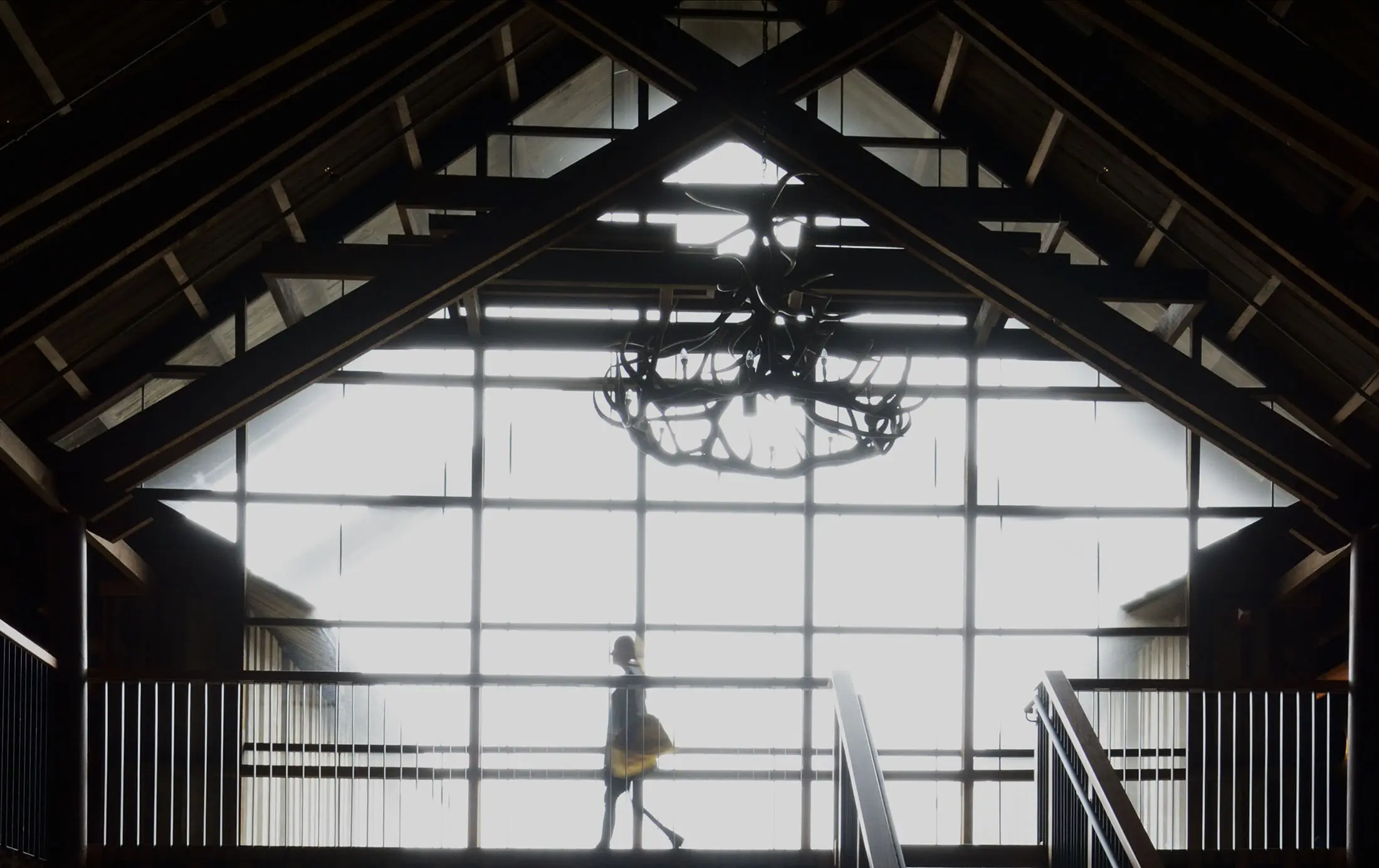 A person with a bag walks across a bridge inside a large building with a high ceiling. The silhouette is visible against a backlit grid of windows. The dark wooden beams and a central chandelier with antler-like designs are prominent in the interior architecture.