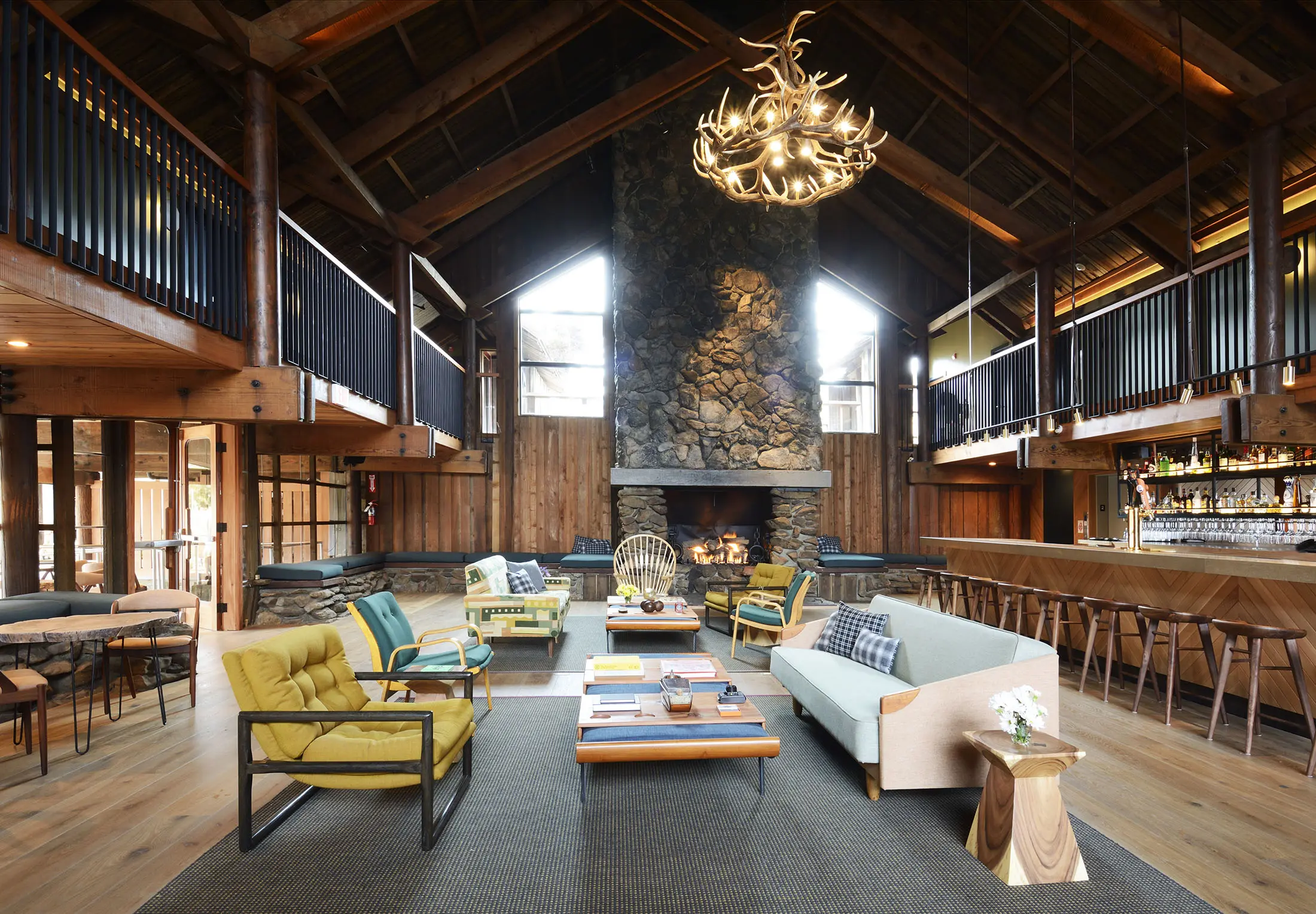 Spacious lodge interior with high wooden ceilings, a central stone fireplace, and modern furniture. Includes sofas, armchairs, and a wooden coffee table on a large rug. A bar with stools lines the right side. An antler chandelier hangs above, completing the rustic yet contemporary ambiance.