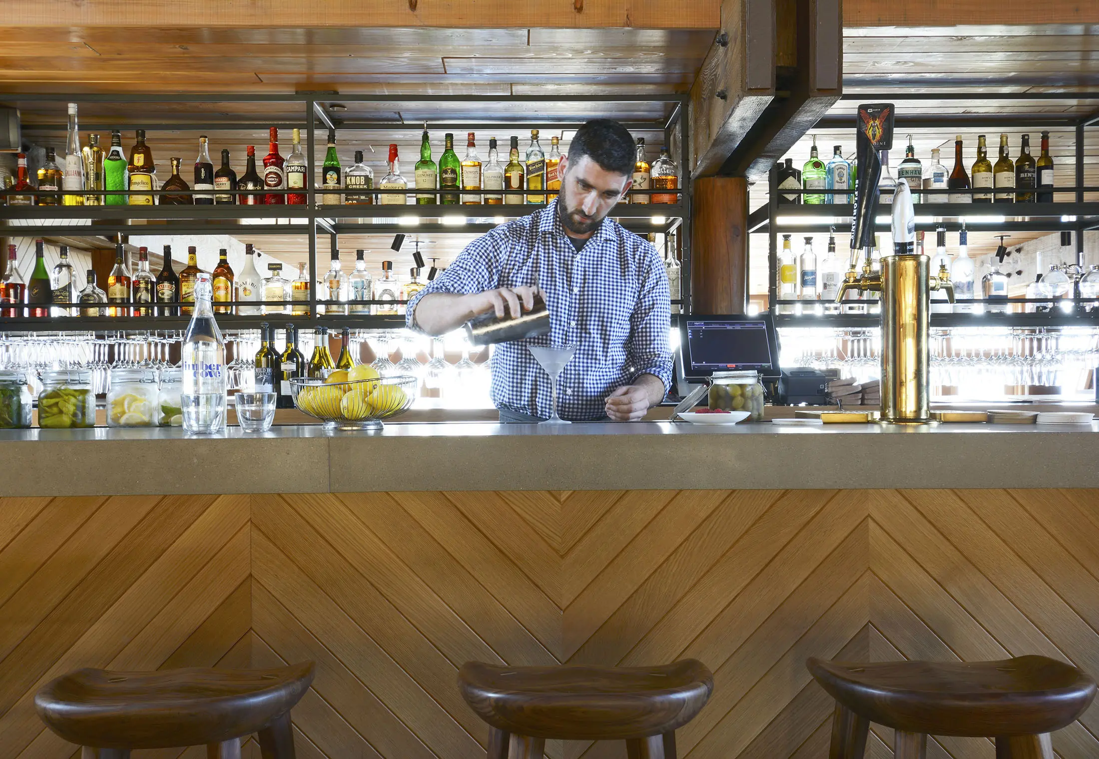 A bartender in a checkered shirt pours a drink from a shaker behind a wooden bar. The bar is stocked with various bottles of liquor on shelves. Lemons, limes, and a glass of water are visible on the counter. Three wooden stools are in the foreground. The setting is cozy and well-lit.