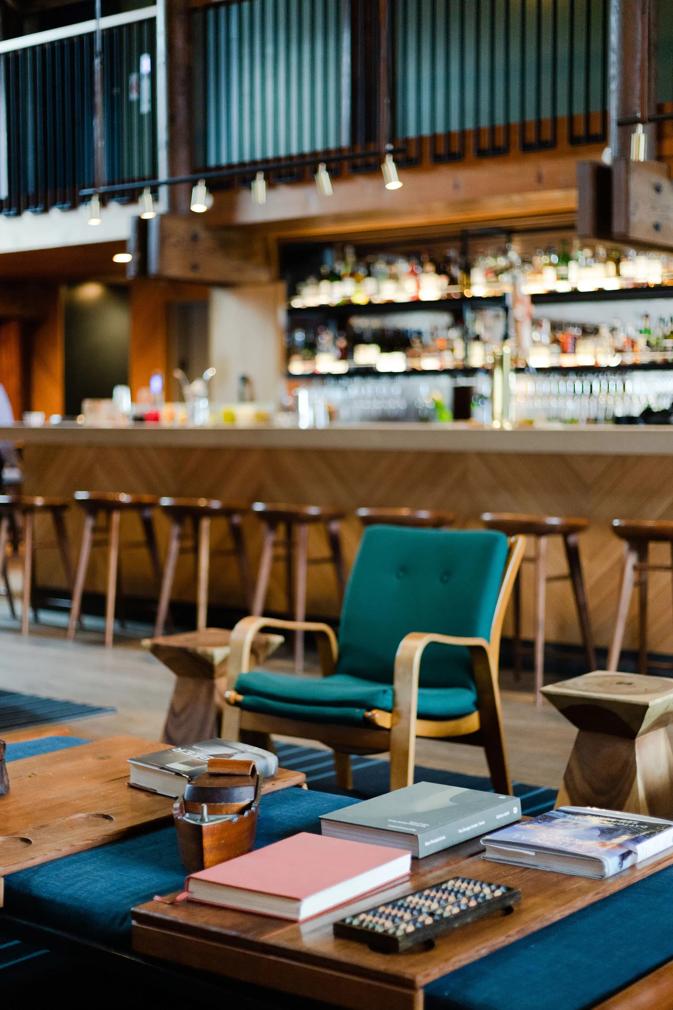 A cozy bar features wooden furniture, a long counter with high stools, and a backdrop of shelves stocked with bottles. In the foreground, a teal armchair and wooden table display books and a basket, creating a warm and inviting atmosphere.