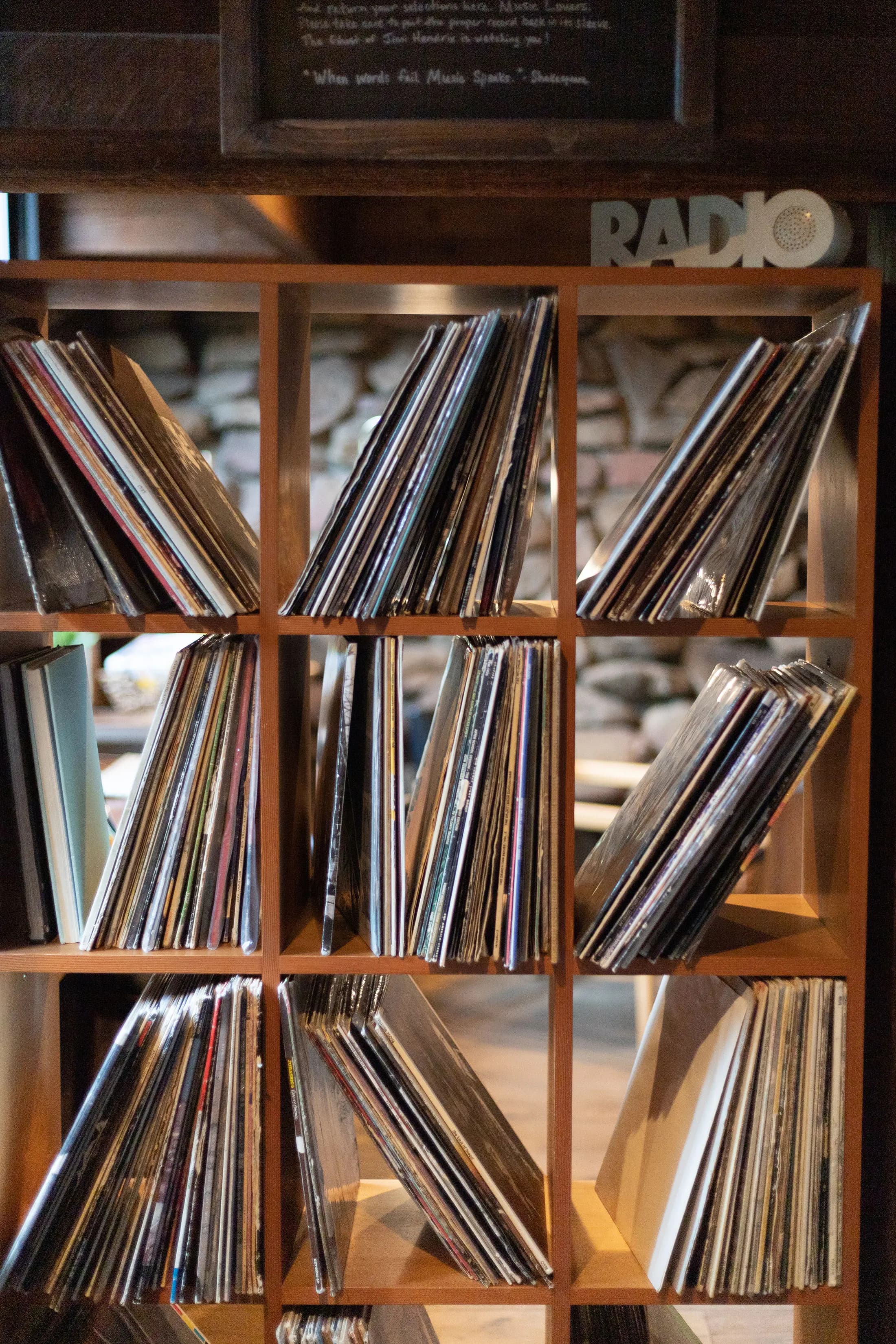 Wooden shelving unit with a grid of compartments filled with vinyl records. A sign with the letters RADIO sits on top of the unit. The shelves are neatly organized, and a stone wall is visible in the background. A menu board is partially seen in the top left corner.