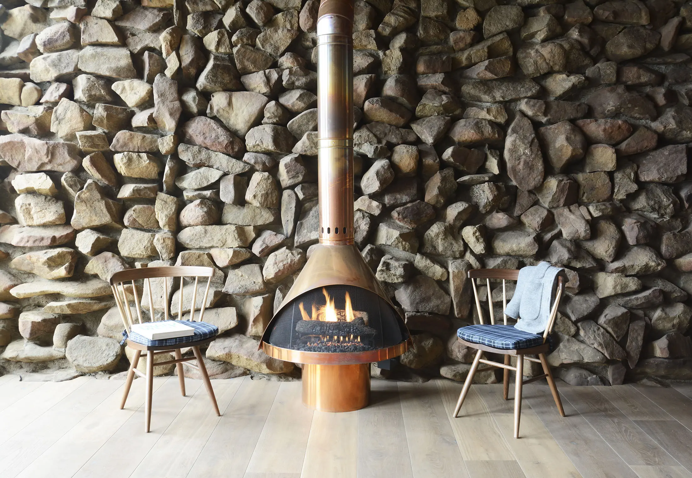 A rustic stone wall features a central copper fireplace with a lit fire. Two wooden chairs with blue plaid cushions flank the fireplace, one on each side. A gray blanket is draped over the right chair. The floor is light wood, and the setting is cozy and inviting.