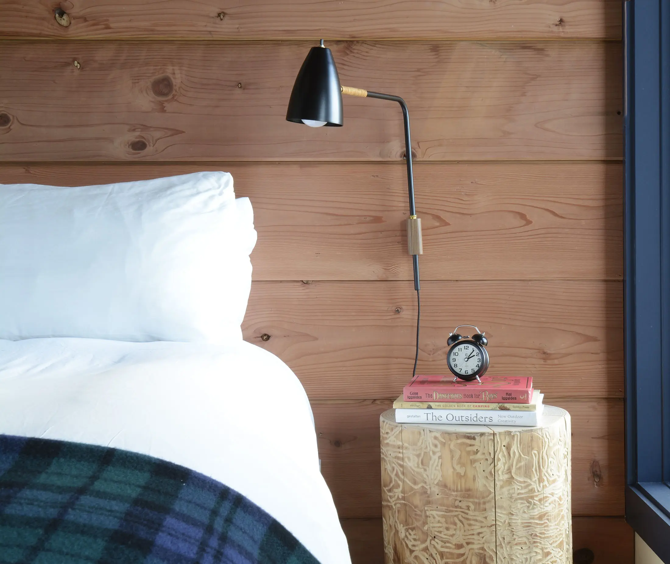 Cozy bedroom corner with a white pillow and bedding on a bed beside a wooden wall. A small round wood side table holds a black alarm clock and two books, The Outsiders and To Kill a Mockingbird. Above them is a modern black wall-mounted lamp. A blue plaid blanket drapes over the bed.