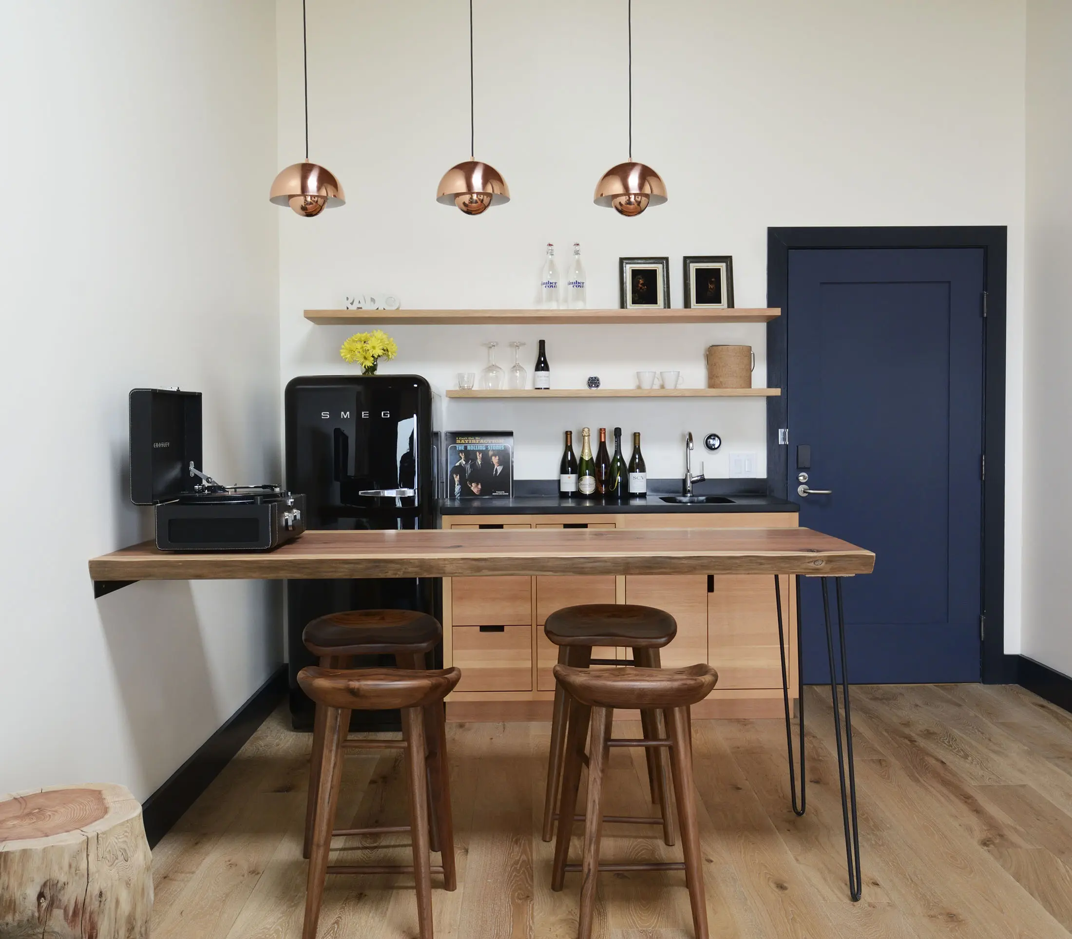 Cozy kitchenette with a wooden bar and three stools. A black SMEG fridge sits on the left. Above, three copper pendant lights hang over the bar. Open shelves hold wine bottles, glasses, and a yellow flower vase. The wall is light-colored with a dark door on the right. Wood floors and a stool.