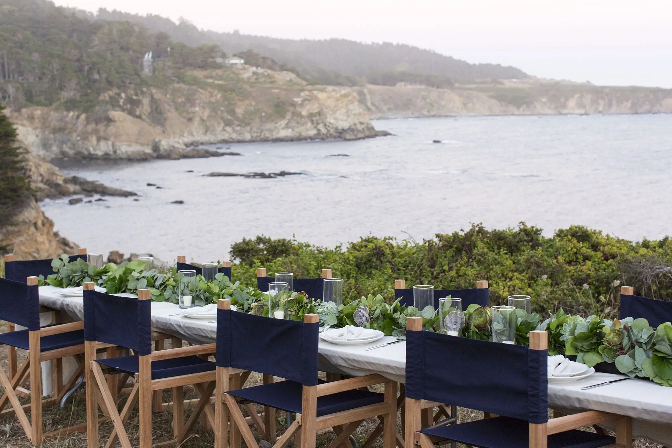 A long outdoor dining table set with plates, glasses, and a green leafy centerpiece overlooks a rocky coastline. Simple wooden chairs with dark fabric seats surround the table. The ocean and cliffs are visible in the background under a cloudy sky.