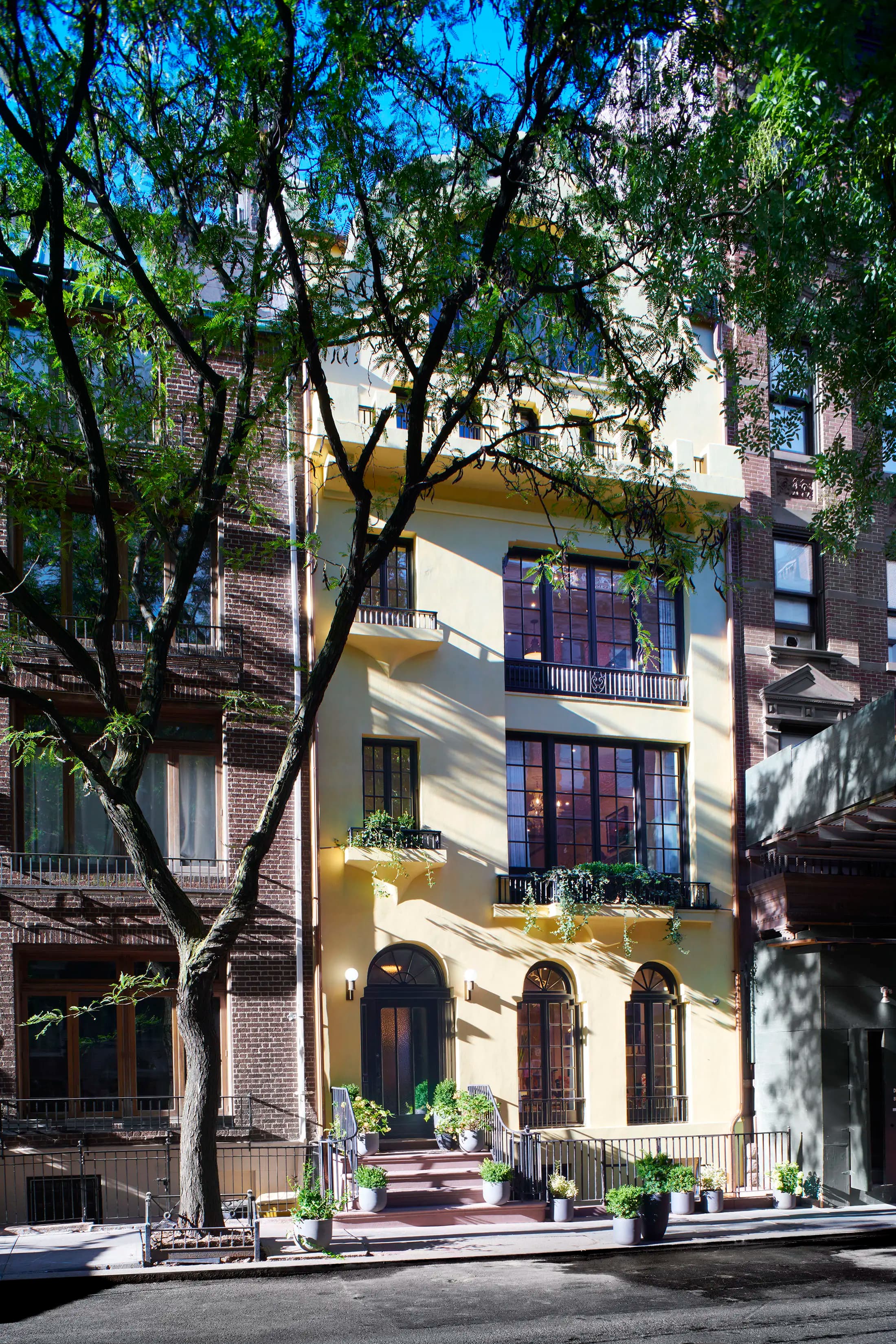 A narrow, yellow building with large arched windows is nestled between two brown brick buildings. Green plants adorn the balconies and a tree with lush foliage shades the street in front. The bright blue sky is visible above, casting subtle shadows on the facade.
